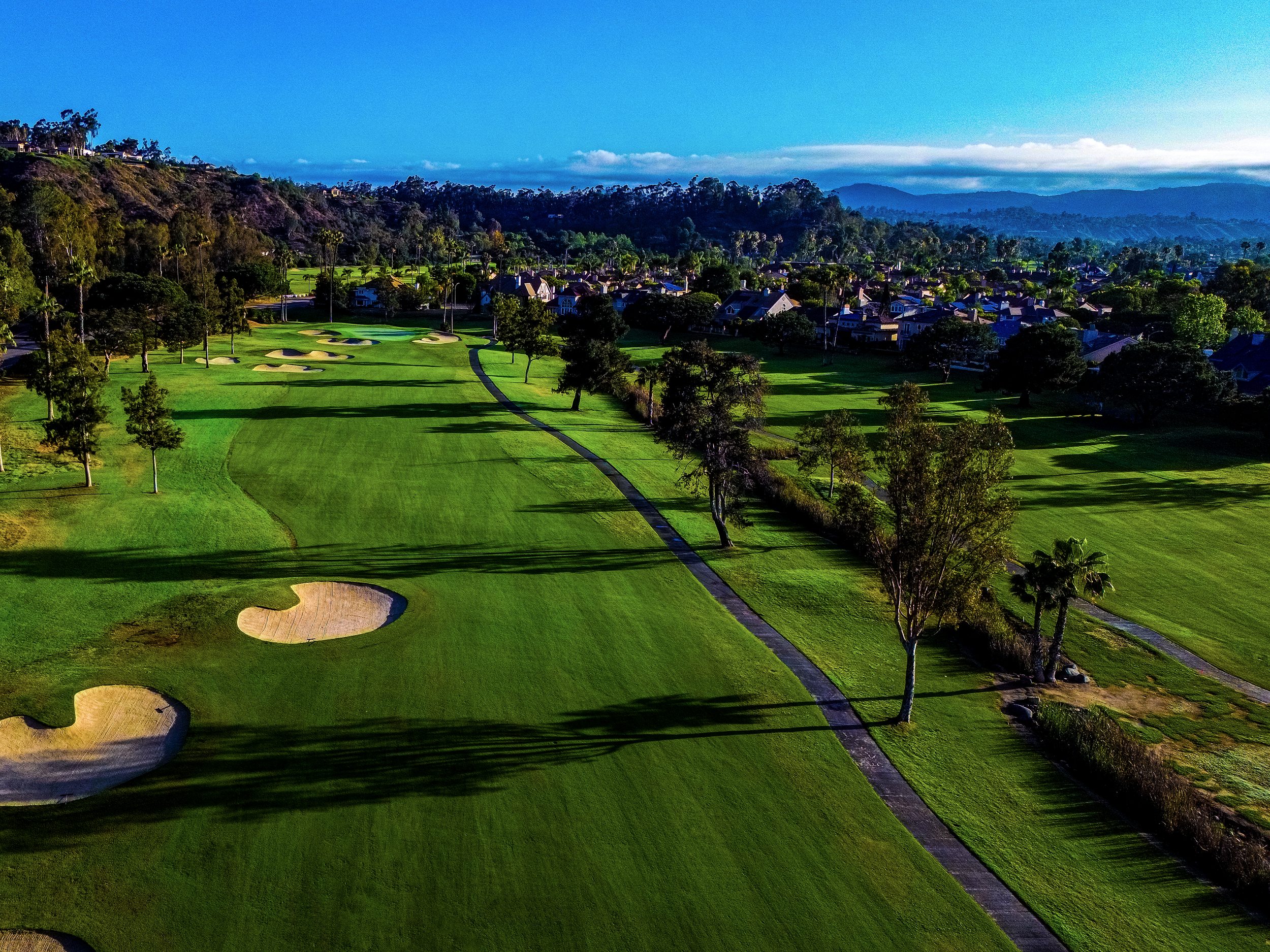 A scenic view of a golf course with sand traps, surrounded by green grass, trees, residential houses, and distant mountains under a clear blue sky.