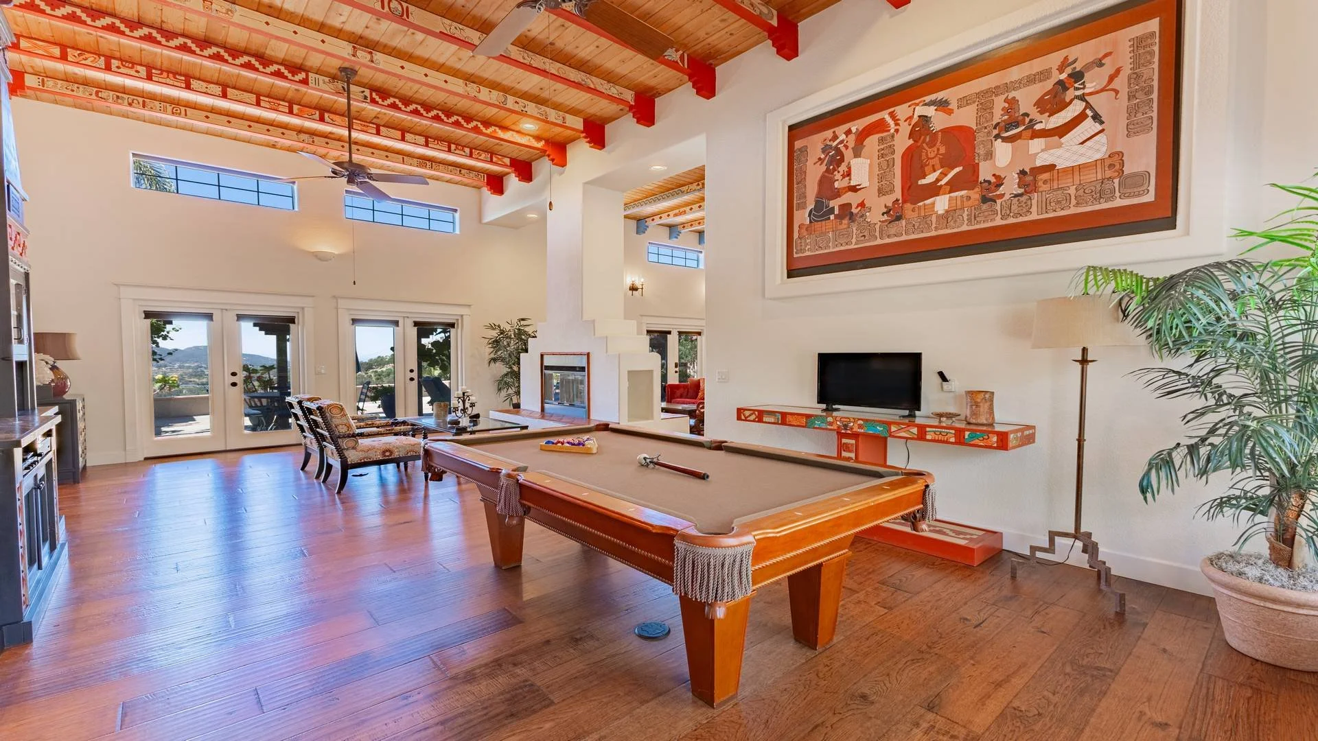 Living room with a pool table in the center, a large artwork on the wall, a TV, large windows, and sliding glass doors leading to an outdoor balcony with a view. Luxury Real Estate Photography in Jamul.