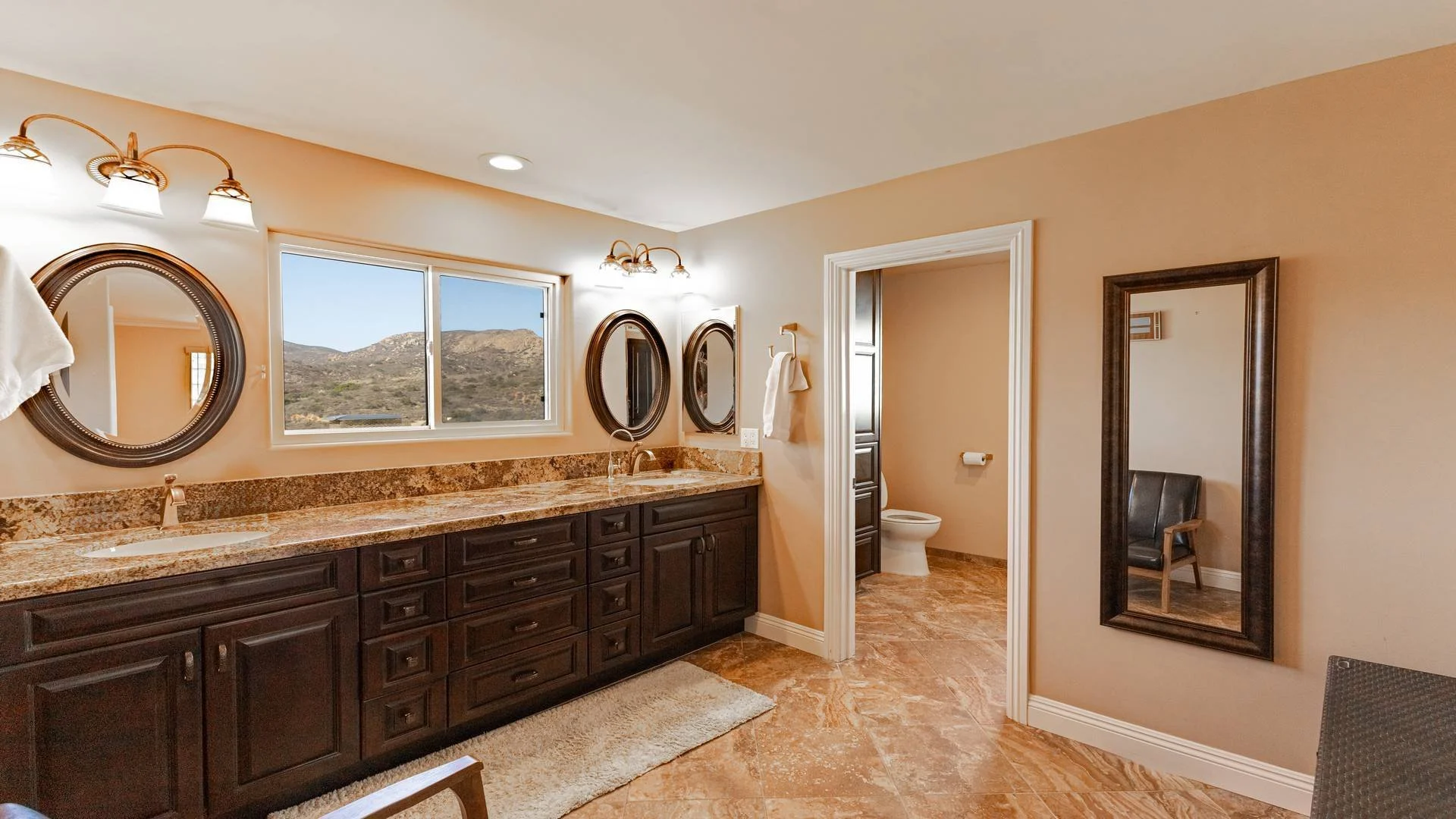 Bathroom with double vanity, granite countertop, oval mirrors, and three-light fixture, view of toilet in separate room, mountain view through window, beige walls, dark wood cabinets, and full-length mirror.