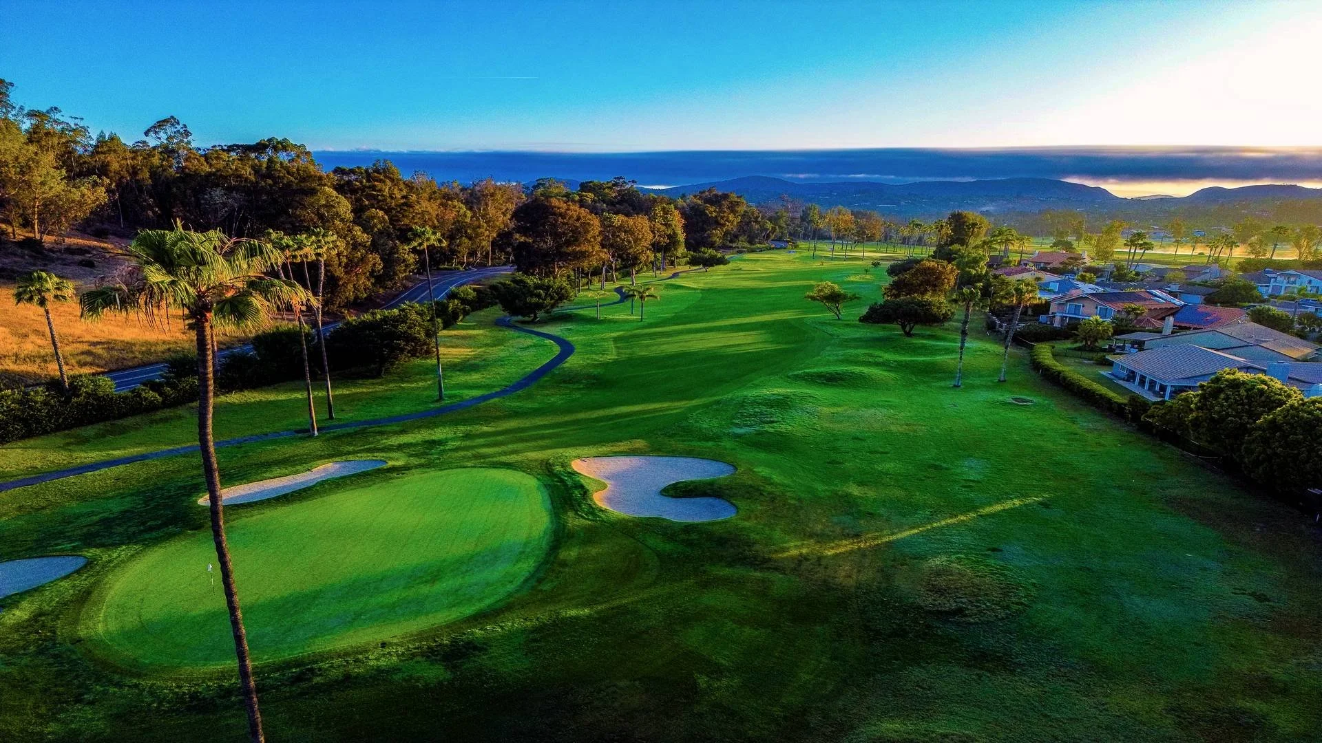 A scenic aerial view of a golf course at sunset, featuring green fairways, sand bunkers, tall palm trees, residential houses along the edge, and distant mountains and ocean under a partly cloudy sky.