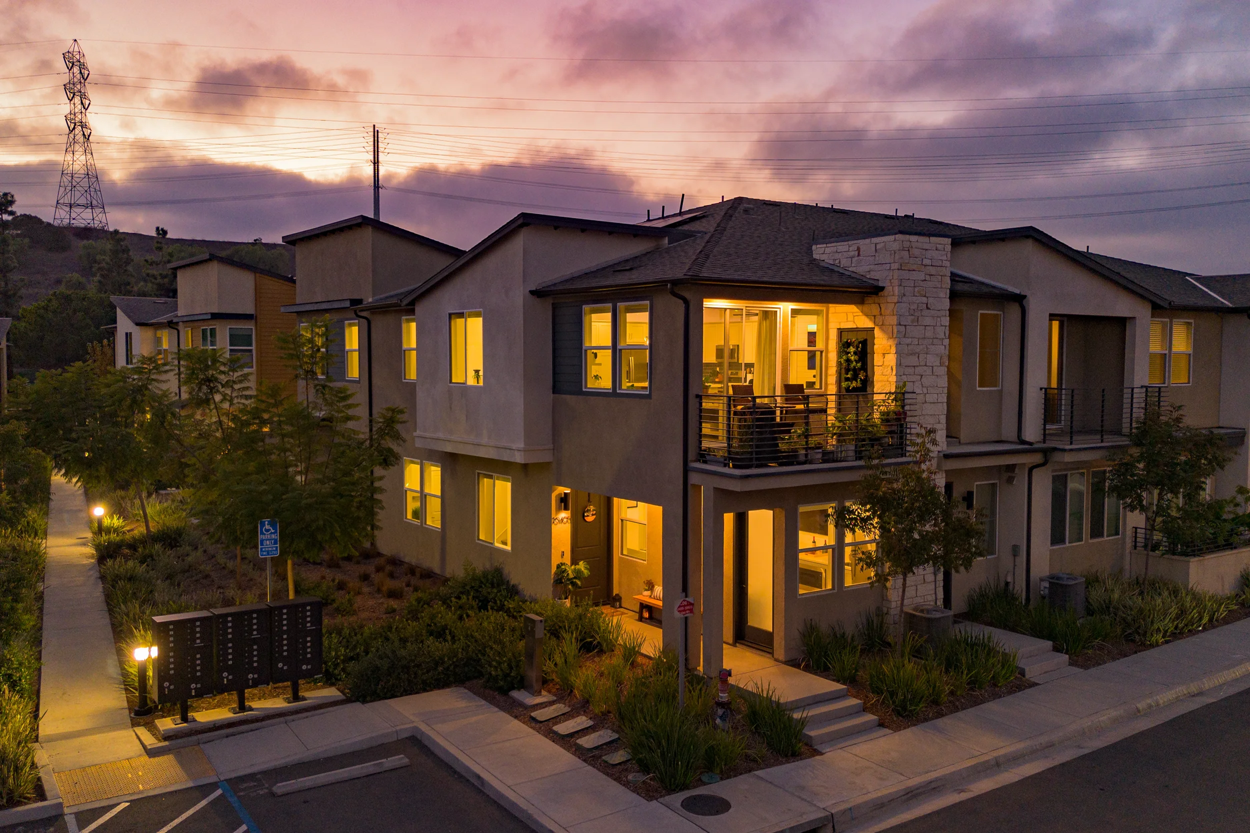 Exterior view of a modern multi-family residential building at dusk with lit windows, balconies, landscaped yard, and surrounding trees.