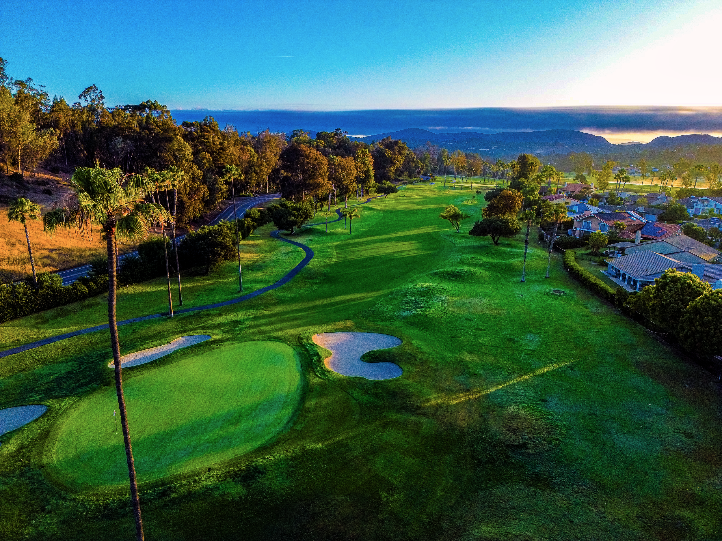 A lush green golf course with sand bunkers, surrounded by tall trees and residential houses, with mountains and a blue sky in the background.