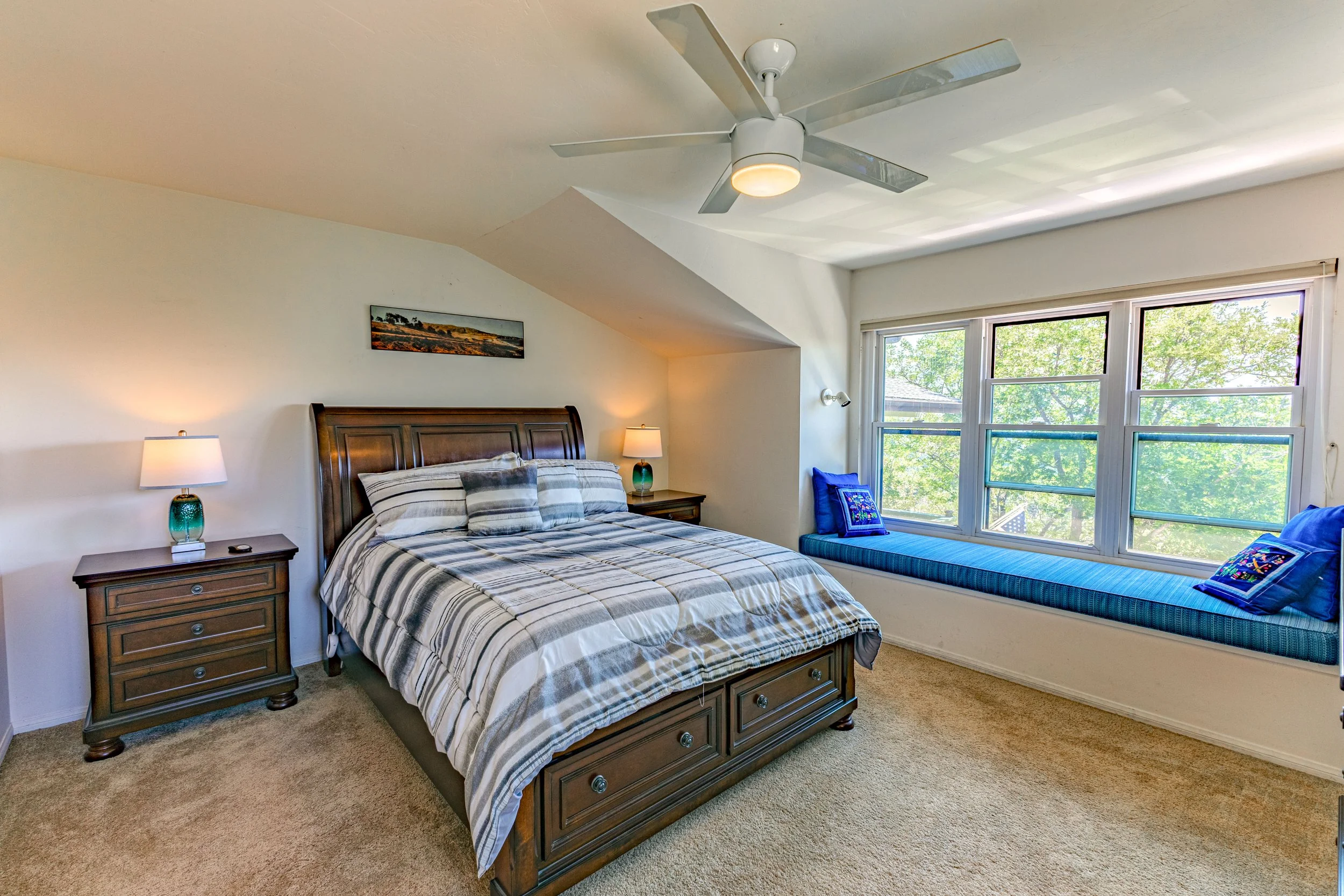 Bedroom with a wooden bedframe, striped bedding, two nightstands with lamps, a window seat with blue cushions and pillows, and a large window showing green trees outside.