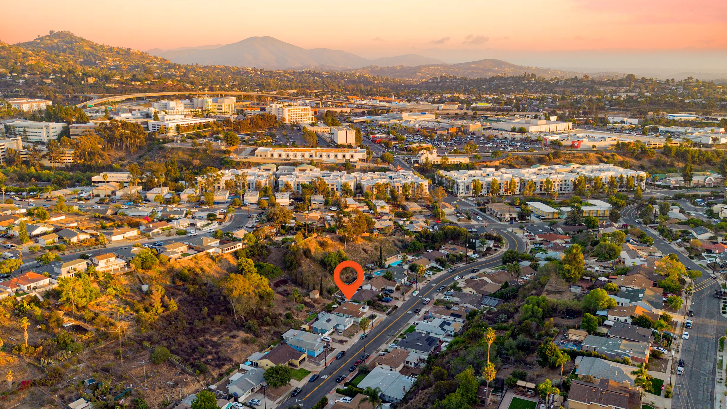 Aerial view of a suburban neighborhood during sunset, showing houses, roads, trees, and a hilly area with a marker indicating a specific house.