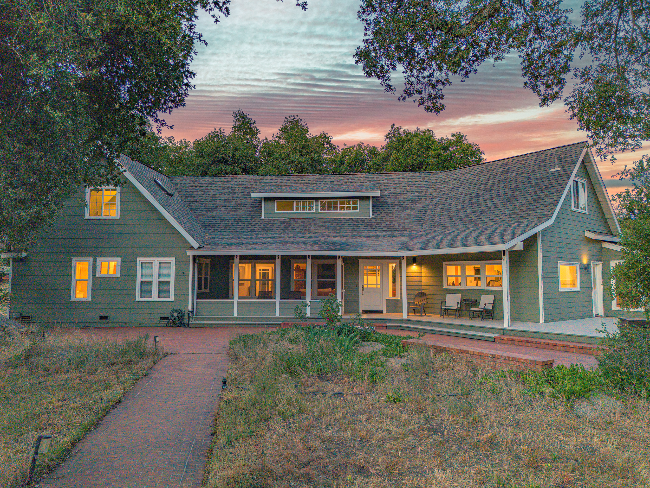 The back of a green house at sunset with a deck, patio chairs, and surrounded by trees and plants.