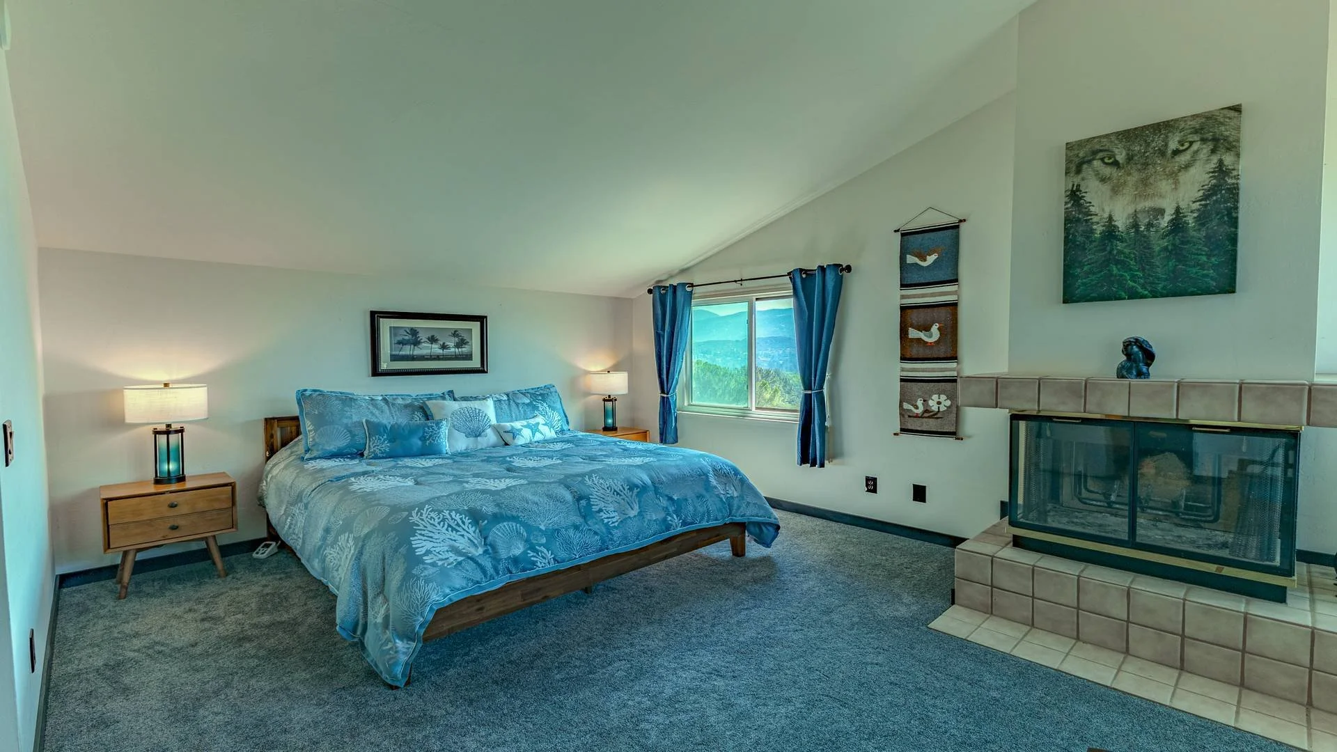 Luxury cabin bedroom with a bed, nightstands, lamps, a window with curtains, and a fireplace with views of a scenic landscape outside the window. Photographed for a San Diego County real estate listing. 