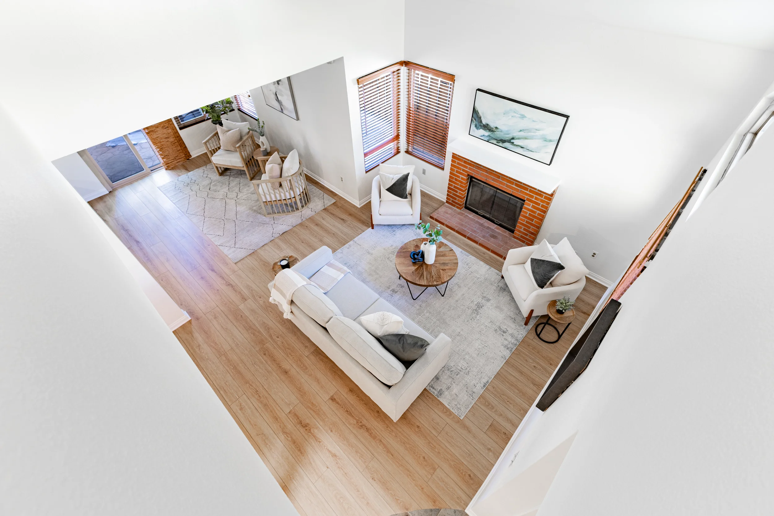 A top-down view of a living room with a white sofa, two white armchairs, and a round wooden coffee table on a light gray rug. There is a brick fireplace with a framed painting above it, and windows with wooden blinds. The room has light wood floors, 