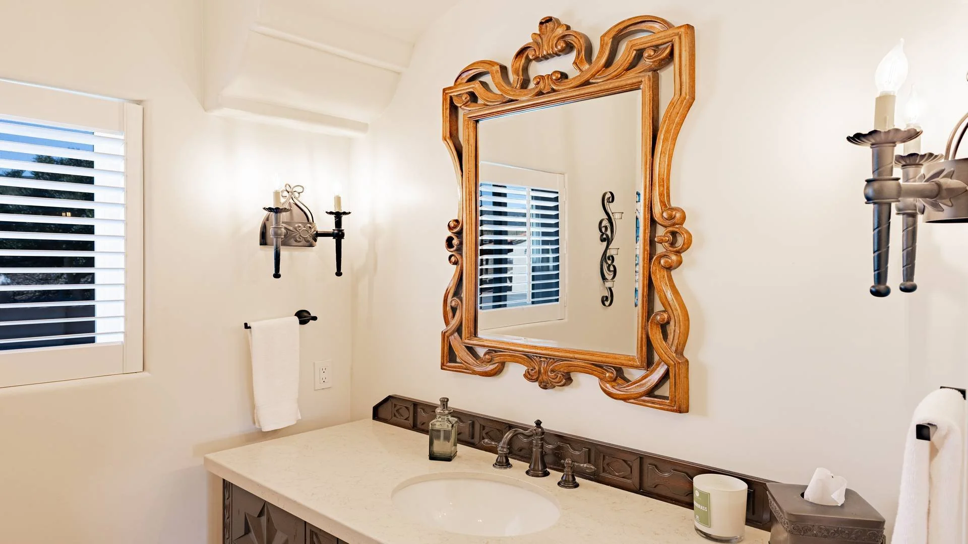 Bathroom vanity with a large decorative wooden framed mirror, black sconces with white candles, a window with white shutters, and a beige countertop with bathroom items.