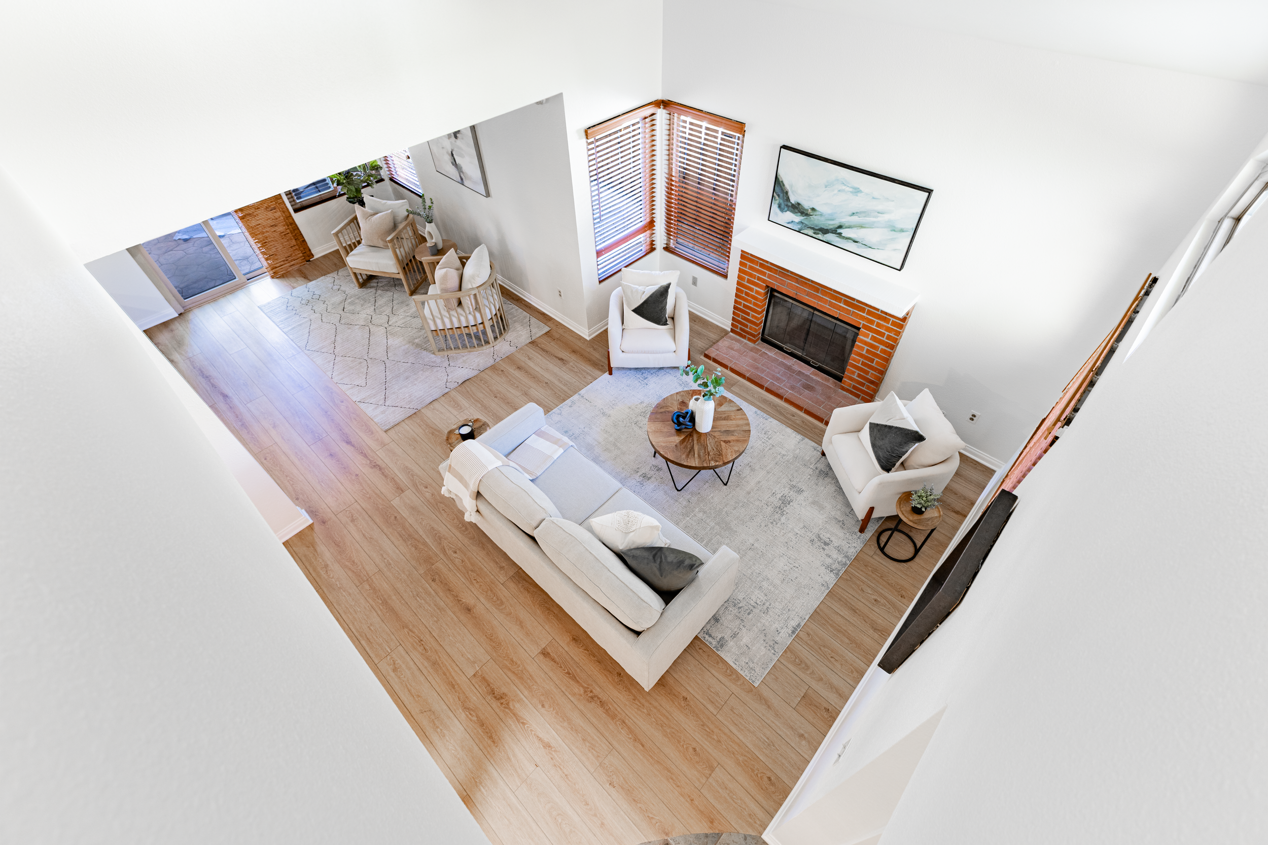A view of a living room with a white sofa, two white armchairs, a round wooden coffee table, a fireplace with a brick surround, and large windows with wooden blinds, from an elevated perspective.