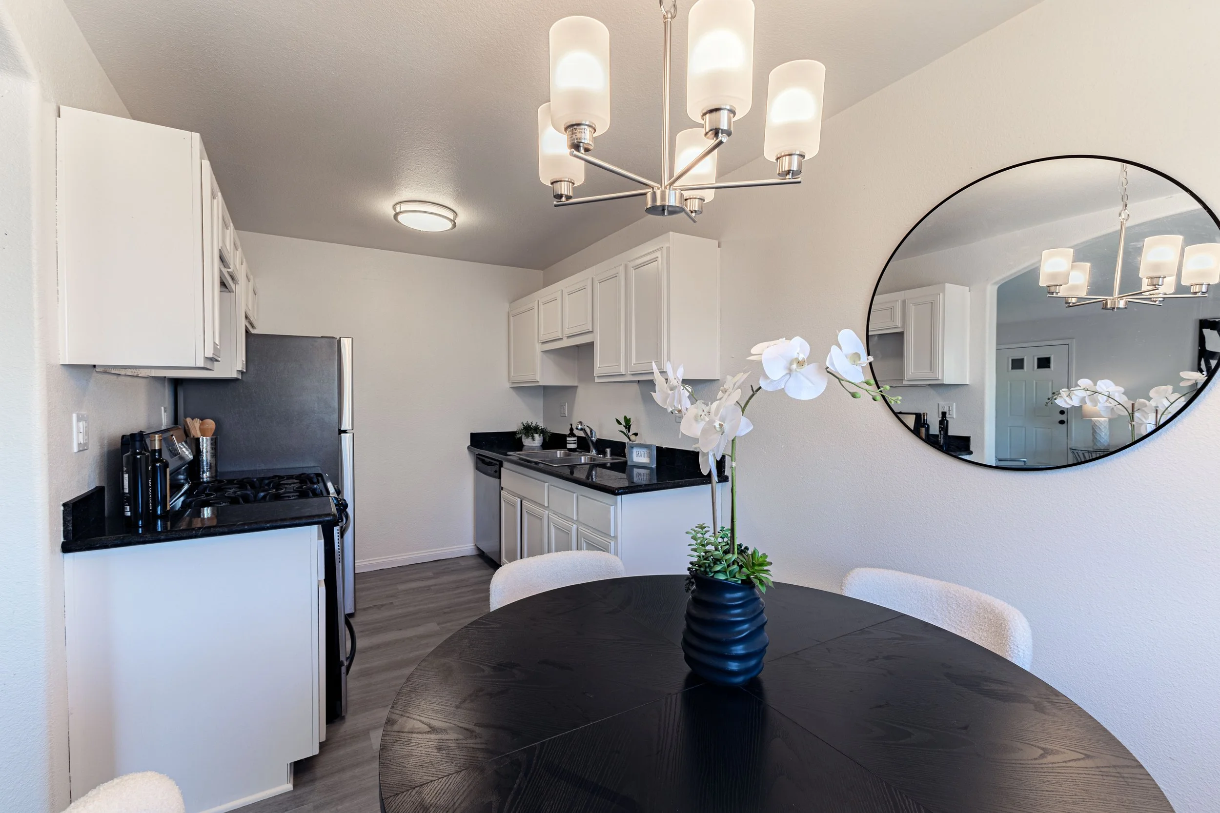 Kitchen and dining area with a black round table, white chairs, a black vase with white orchids, white cabinets, black countertops, a stainless steel refrigerator, and a round wall mirror.