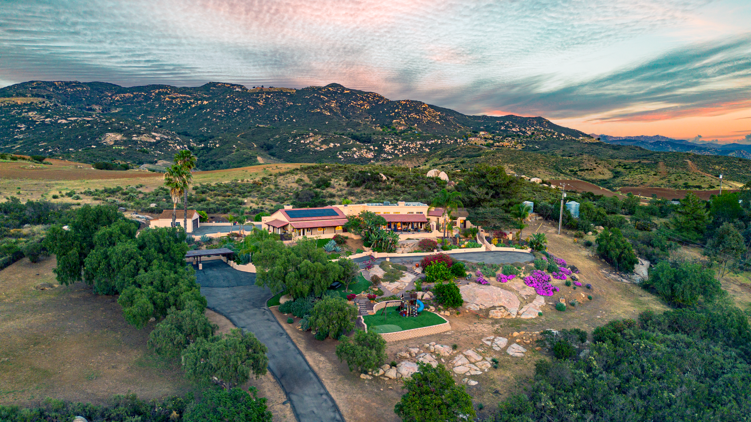 A large estate in a hilly, semi-arid landscape during sunset with a main house, lush greenery, colorful flowering bushes, and a winding driveway.