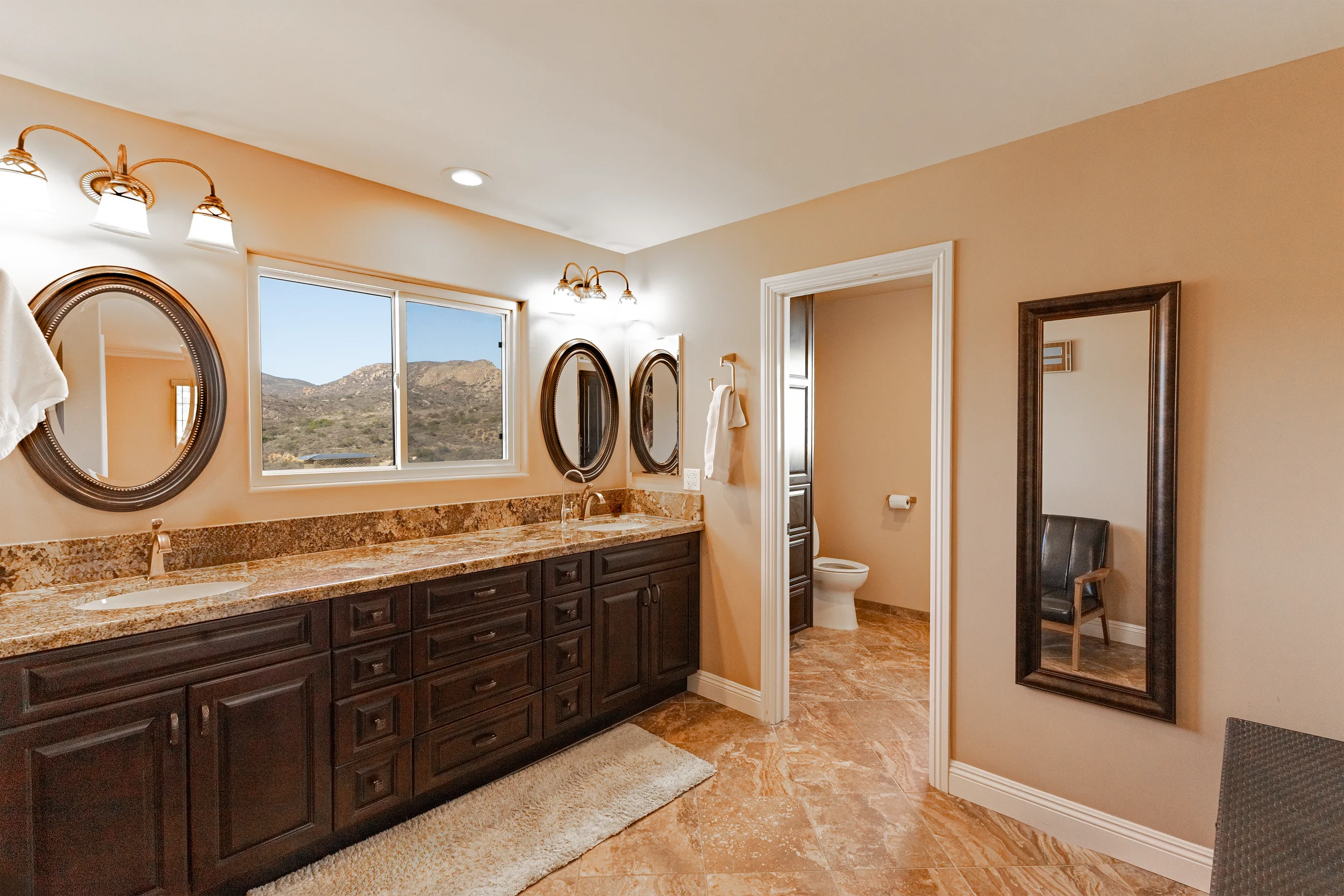 Bathroom with dual sinks and granite countertops, oval mirrors, window with mountain view, a toilet in a separate room, and a full-length mirror on the wall.