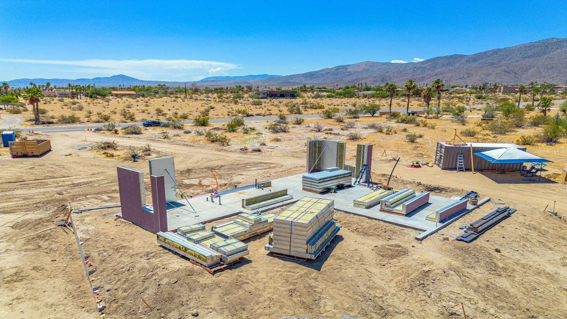 Construction site in a desert landscape with building foundations, construction materials, and a few small structures, with mountains and a clear blue sky in the background.