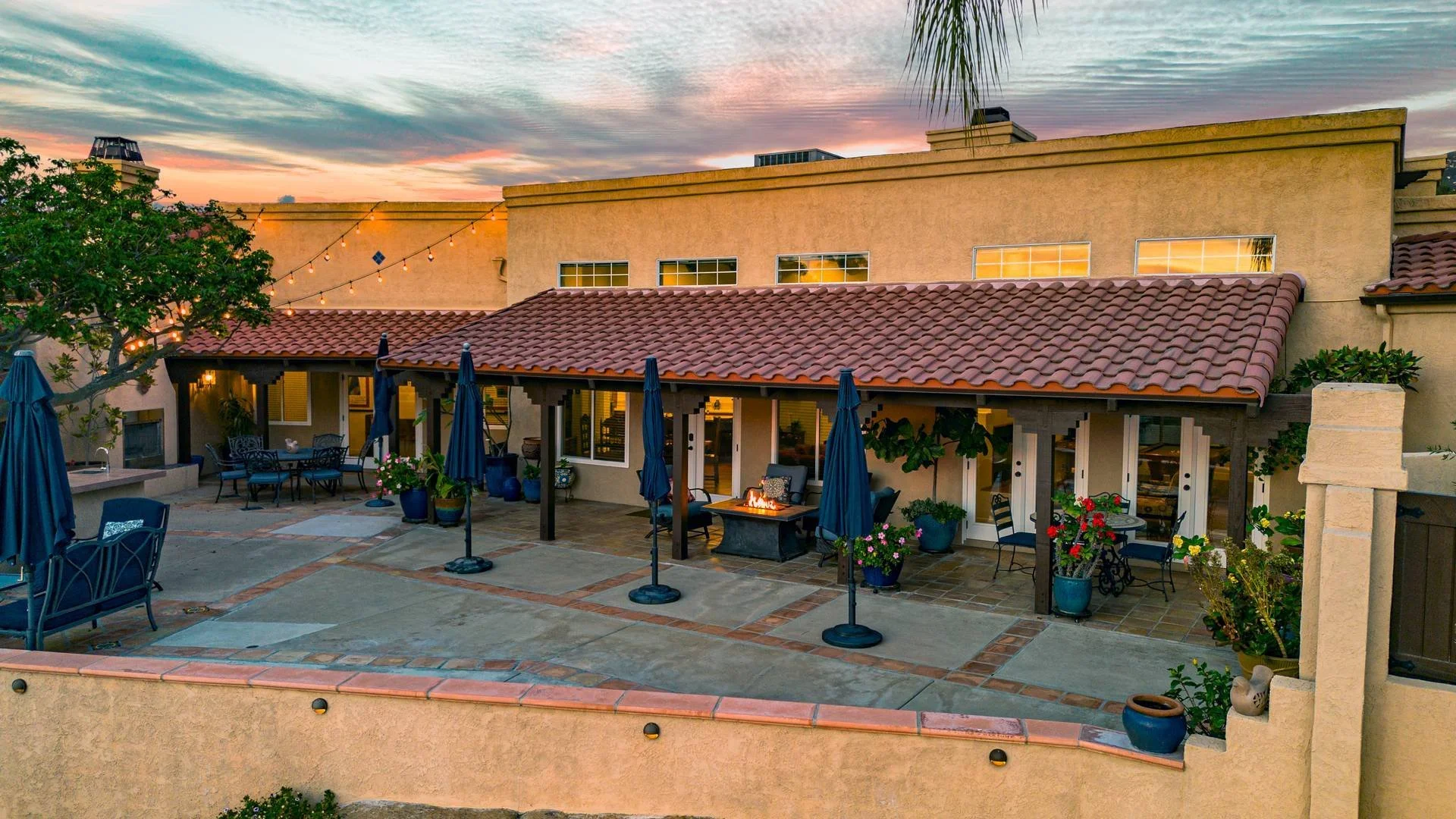 Backyard patio with outdoor furniture, string lights, potted plants, and a firepit at sunset, in front of a beige stucco house with a red tile roof.