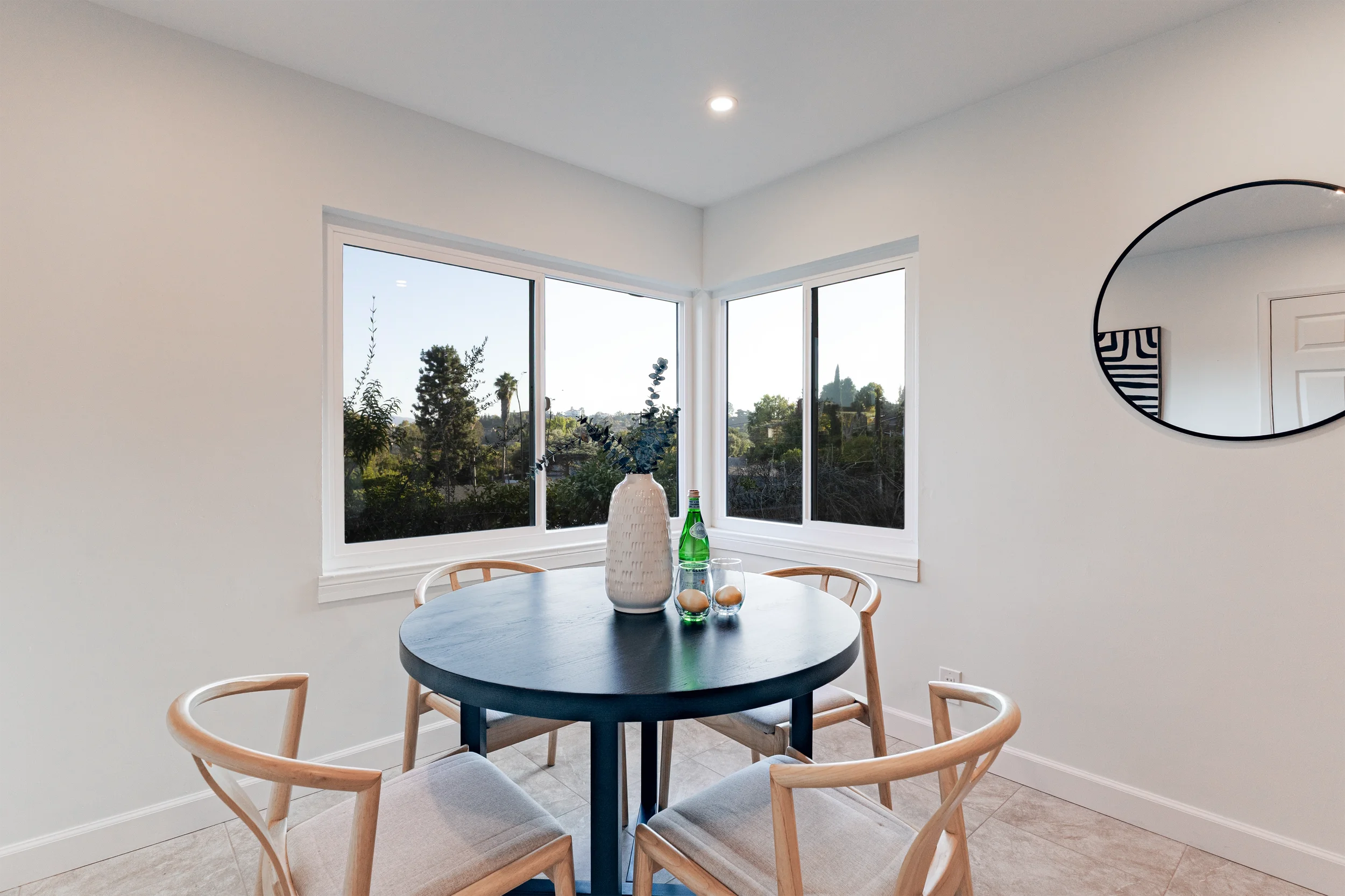 Dining area with a round black table, four cream-colored chairs, a white vase with blue flowers, a glass bottle, and two glasses on the table, with large windows showing outdoor greenery.