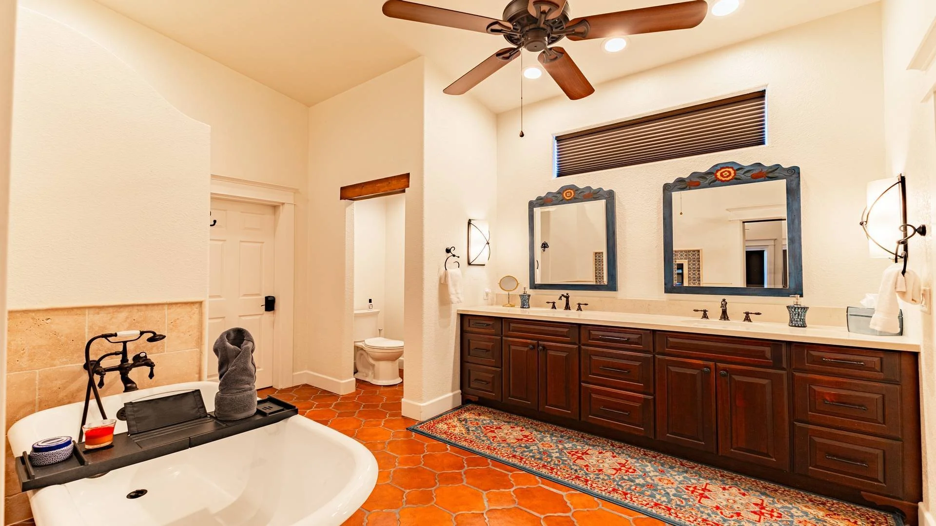 Bathroom with a large double vanity with two mirrors, a bathtub with a black faucet, terracotta tiles on the floor, and a ceiling fan.