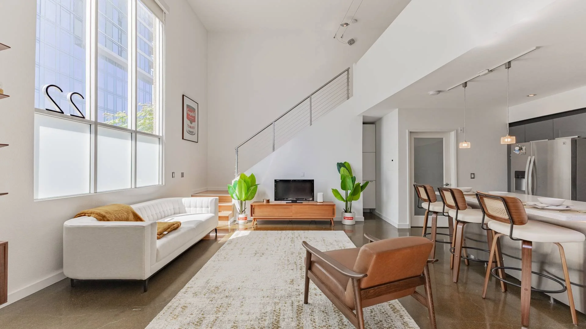 Modern open-concept living room with white walls, large windows, a white sofa with a brown blanket, green potted plants, and a dining area with wooden chairs and a long white table. Interior Photography in downtown San Diego. 