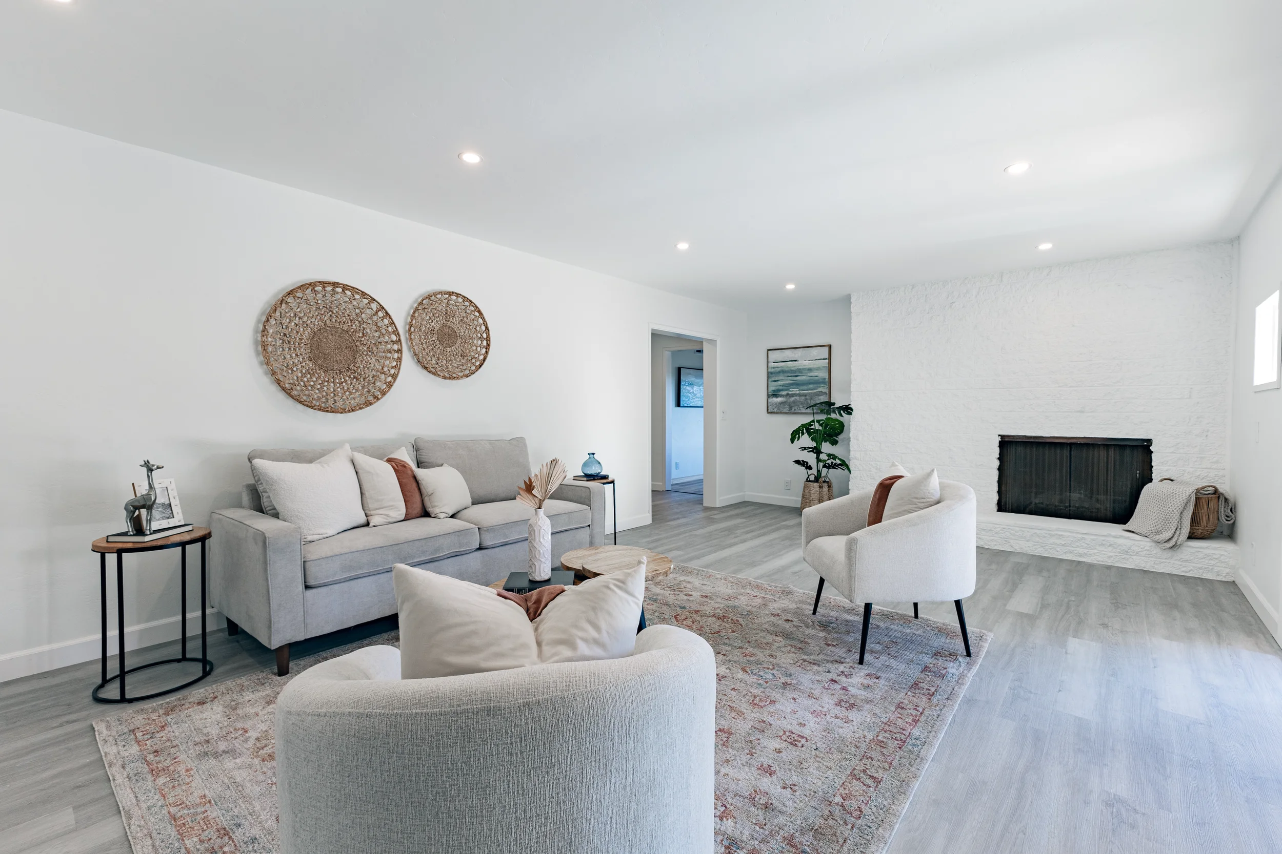 Bright living room with light gray sofa, white armchair, and beige area rug. White walls, decorative wicker wall hangings, fireplace, and potted plant enhance the space. Recessed ceiling lights and minimal decor complete the look.