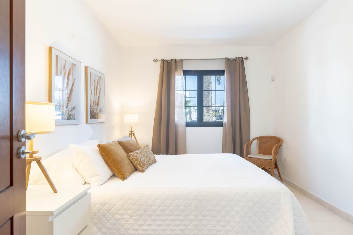 Cuarto con cama grande, almohadas marrones y decoraciones naturales, cortinas beige, ventana con vista a palmeras, lámparas de mesa, silla de mimbre, paredes blancas y piso claro.