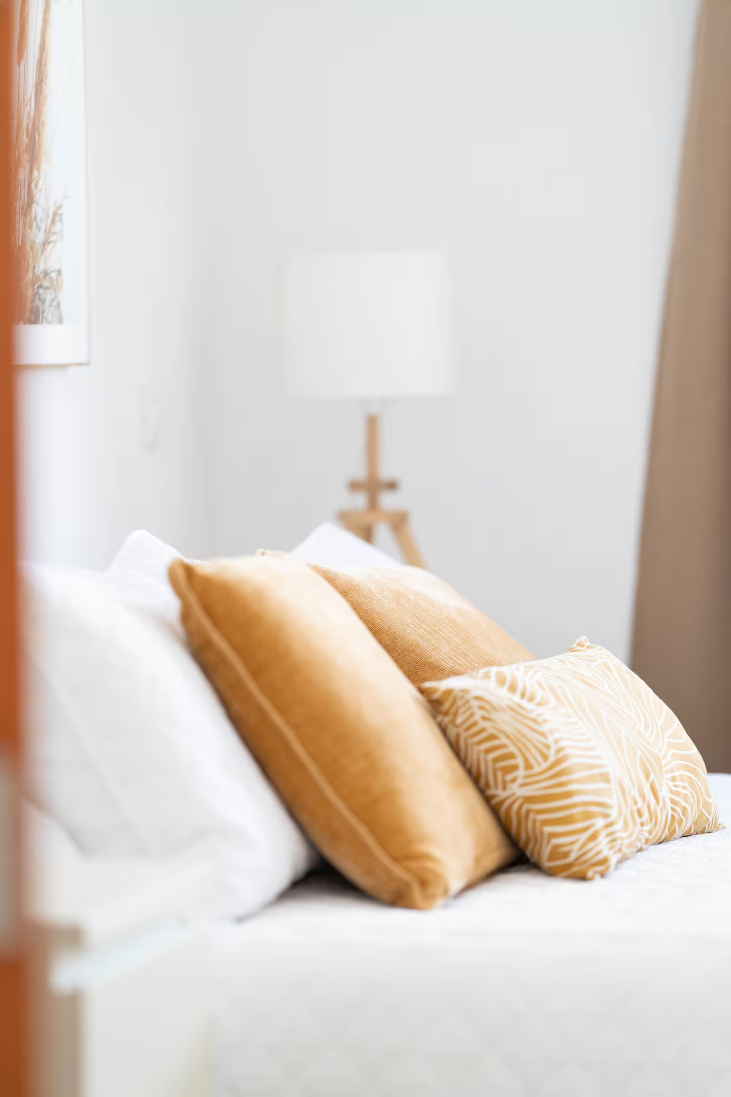 Cojines en tonos dorados y beige sobre una cama blanca, con lámpara de pie de madera y cortinas beige en fondo.