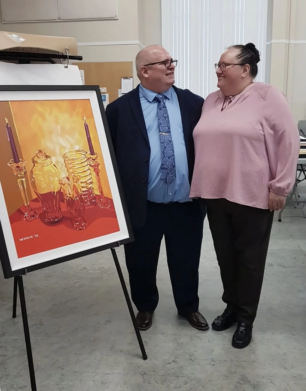 Janet and David Weirmeir at an art show, that he had at Cooke's Presbyterian Church, in Chilliwack, BC, Monday, December 8th, 2025. Beside them is a painting, titled "Glassware and Crystal" on an easel.