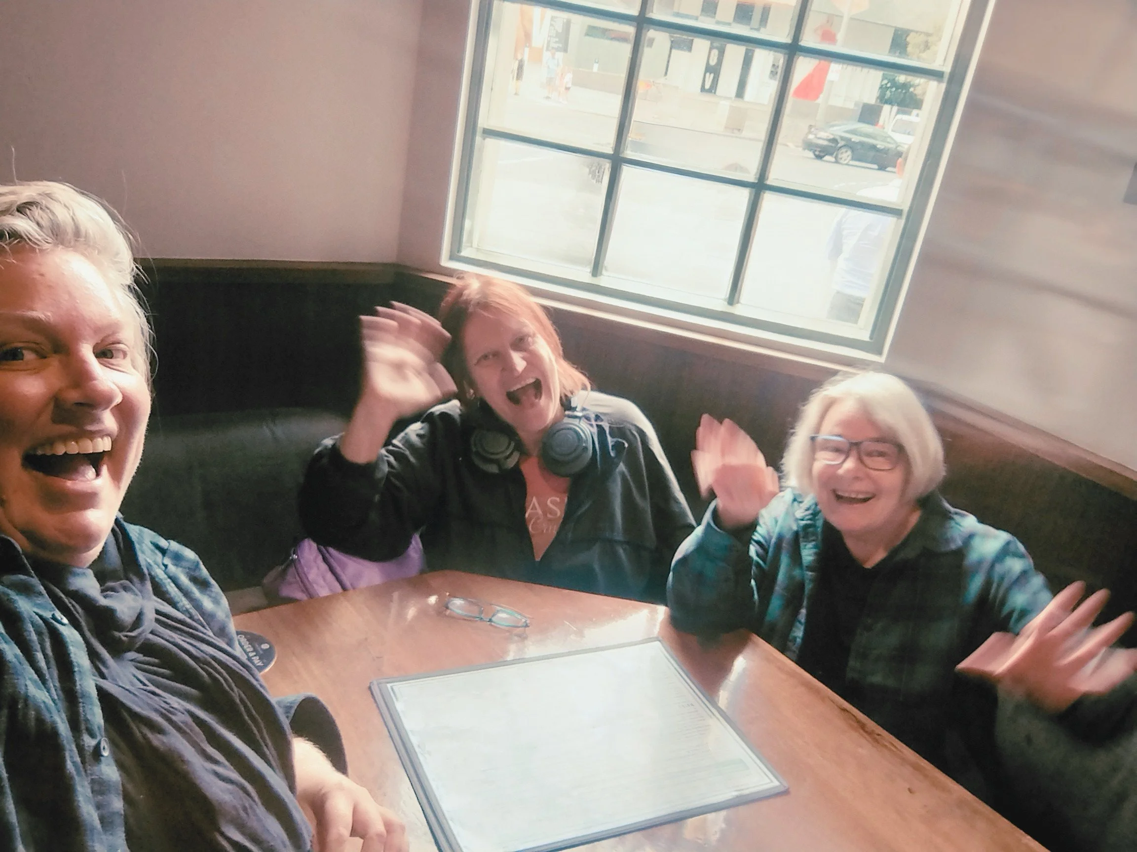 Three smiling people sitting at a table, raising their hands and waving, with large windows behind them.