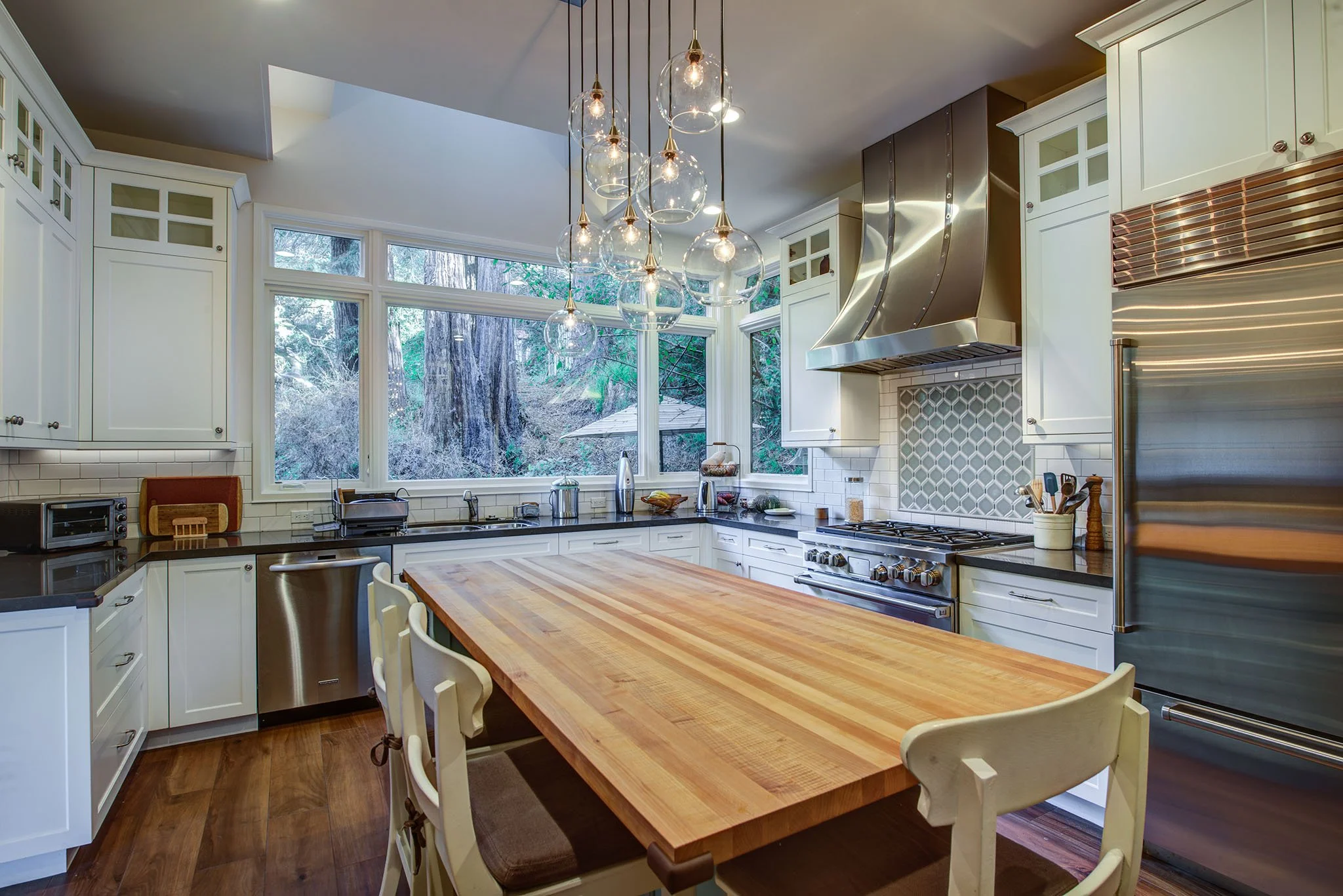 Sunlit kitchen with broad counter seating, white cabinets, tile backsplash, and views of the wooded site, reflecting Levitch craft details in a 1920s inspired remodel.