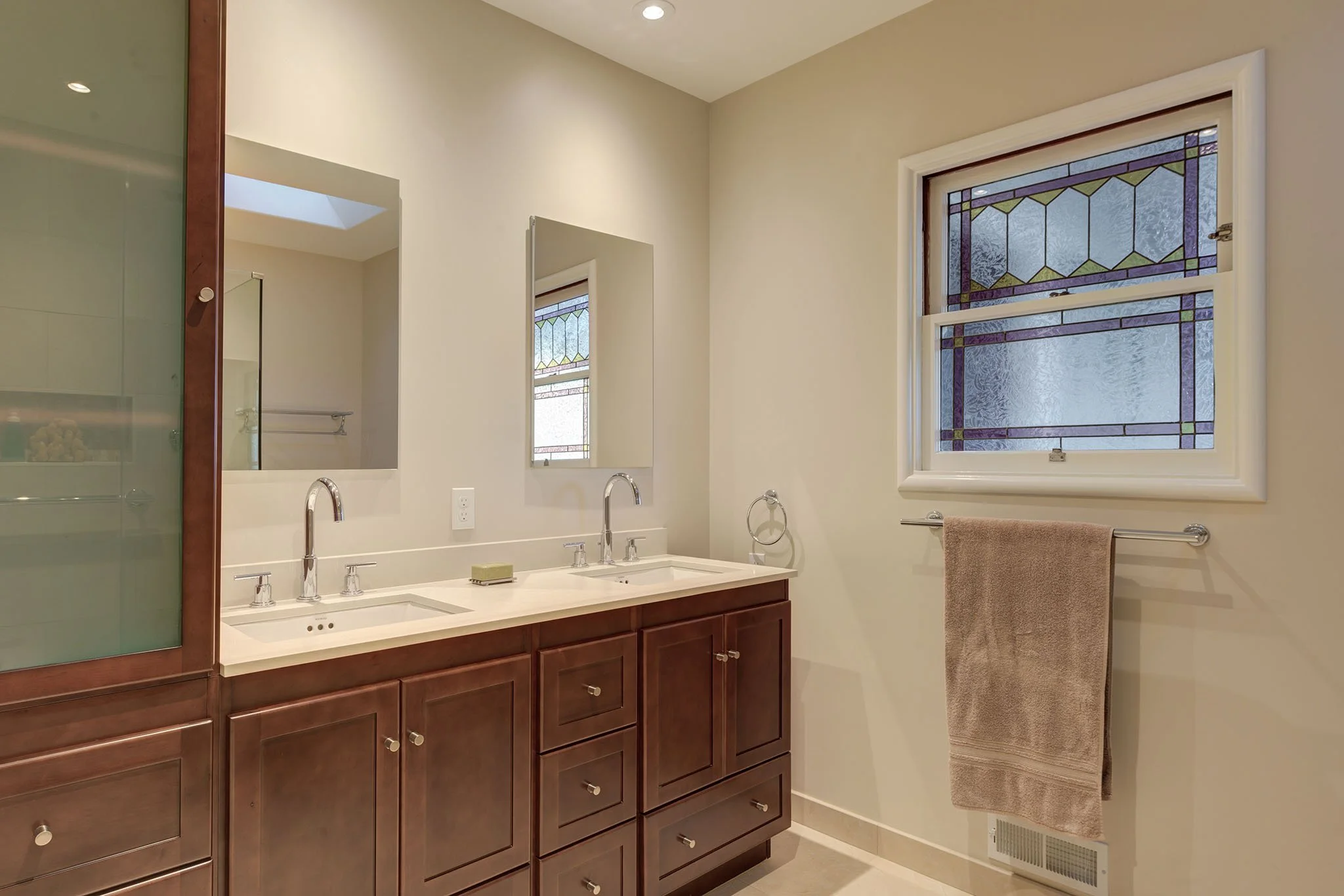 Levitch remodeled primary bathroom with a double sink vanity, white countertop, dark wood cabinetry, chrome faucets, and mirrors beside a stained glass window and towel bar.