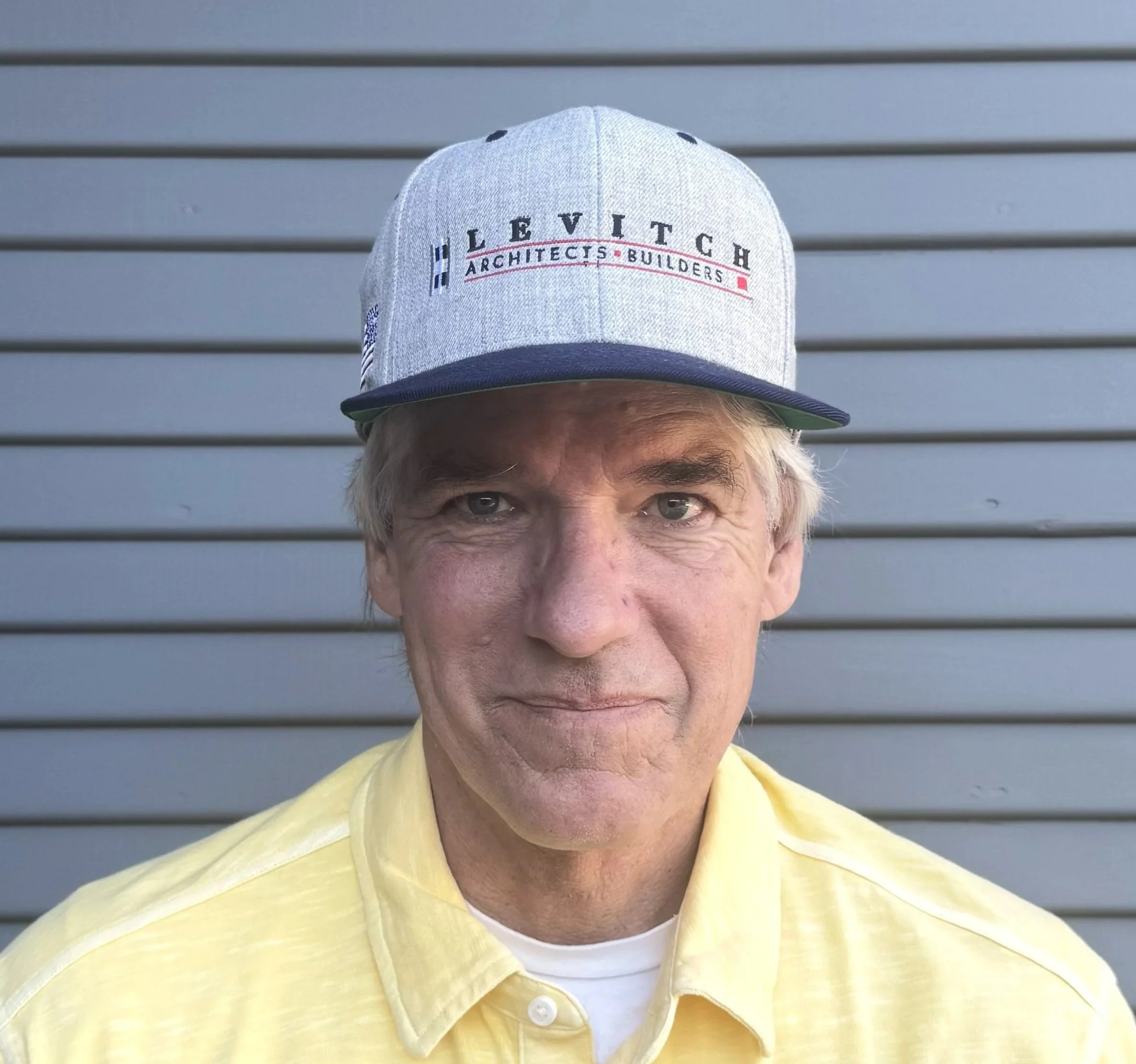 A middle-aged man wearing a grey baseball cap with the words 'Hillevitch Architects Builders' embroidered, and a yellow button-up shirt, standing in front of a grey, horizontal-paneled wall.