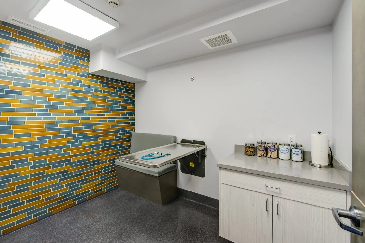 Exam room with stainless pet table and storage cabinets set against a blue and yellow tiled accent wall, adding color while keeping the space bright and hygienic.