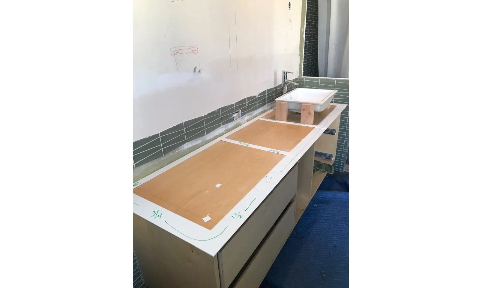 Master Bathroom remodel in progress showing the custom floating Kirei Board vanity under construction, with layout templates for dual sinks and green glass tile installed on the wall.