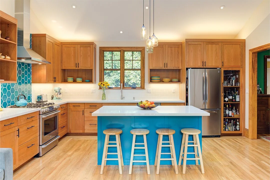 View across the kitchen island toward the cooking area, highlighting turquoise accent tile at the range, warm wood cabinetry, and pendant lights beneath a vaulted ceiling.