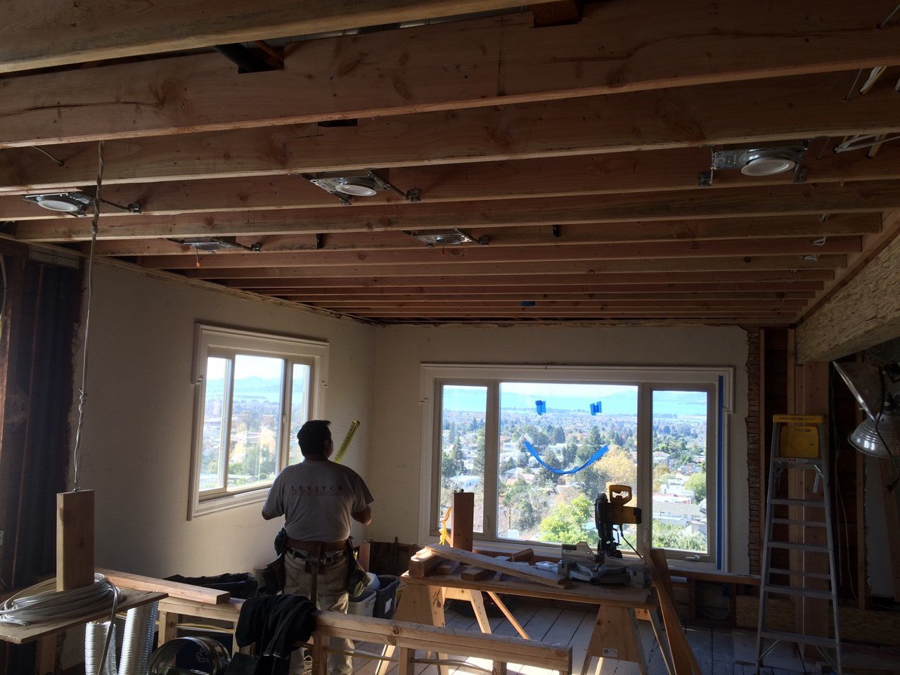 Ceiling opened to expose framing for updated lighting in the open plan living space, with a carpenter at work and wide windows framing Bay views in the distance.