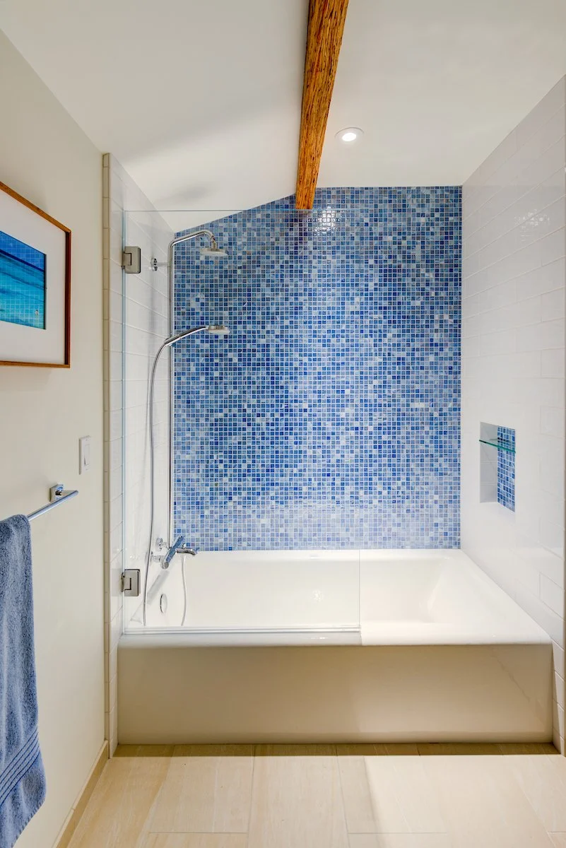 Bright, modern bathroom with a tub-shower combo featuring a blue mosaic tile accent wall, white subway tile surrounds, glass partition, and warm wood beam detail for a fresh coastal-inspired aesthetic.