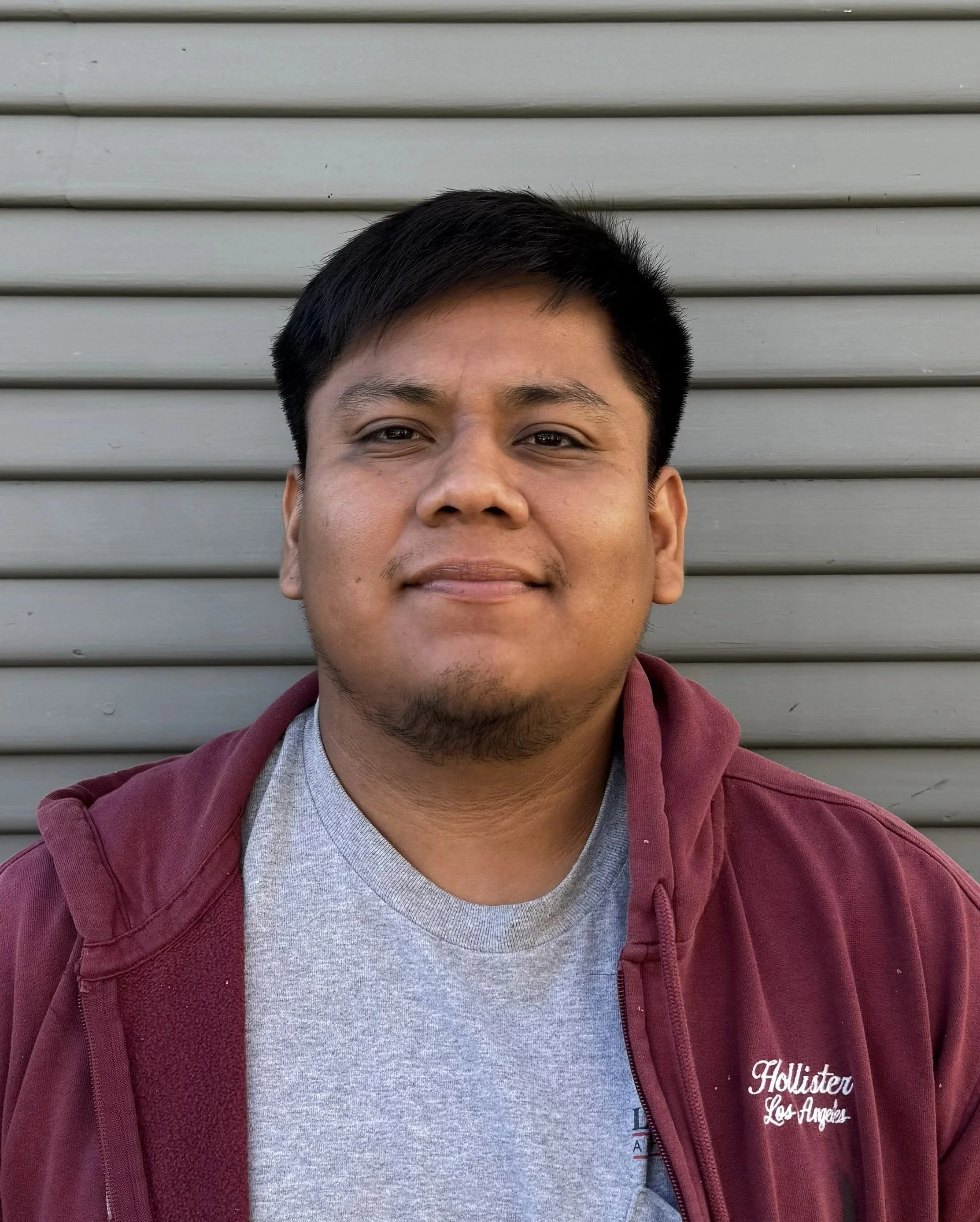 A young man with short black hair and light brown skin, wearing a gray T-shirt and maroon hoodie, standing in front of a gray metal shutter.