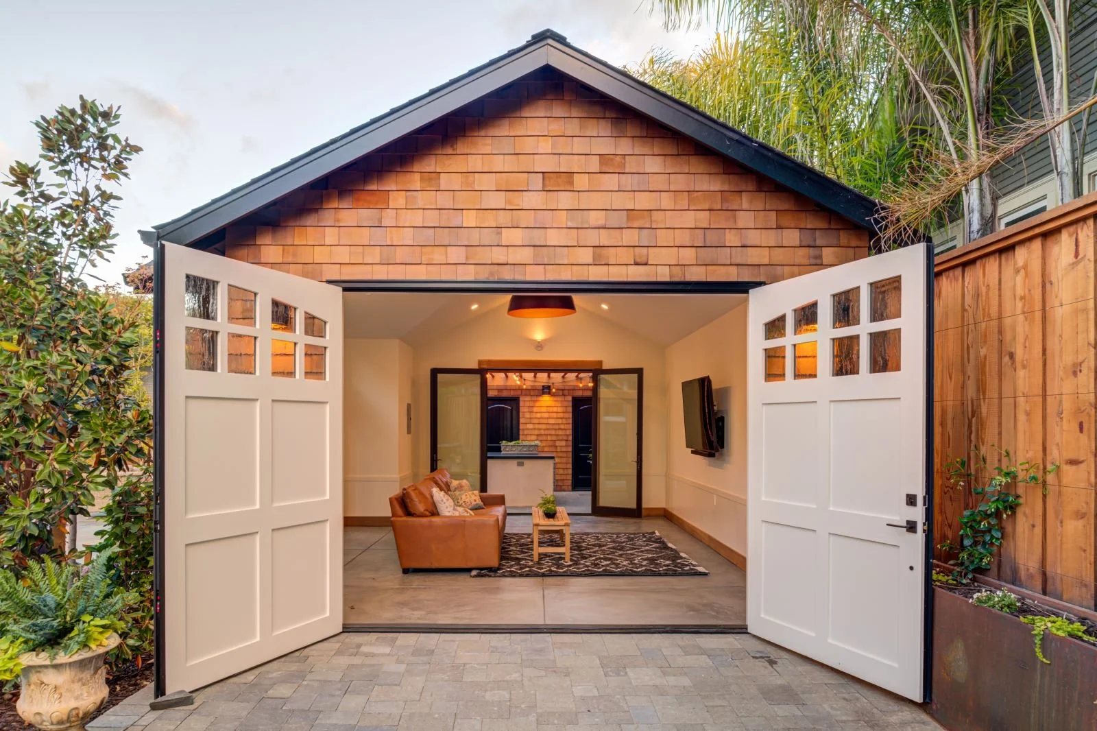 With the doors fully opened, the accessory structure transforms into an indoor–outdoor pavilion, where cedar shingle cladding, painted wood doors, and polished concrete floors support entertaining, work, or relaxation.