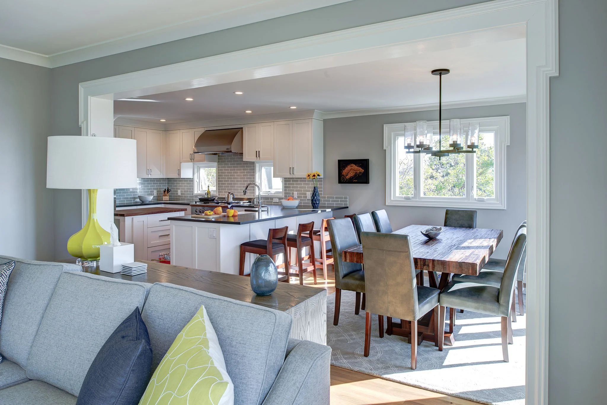 Open plan living, dining, and kitchen remodel with a broad cased opening, gray and white finishes, and a long dining table beside bright windows. Spaces connect easily for relaxed entertaining.