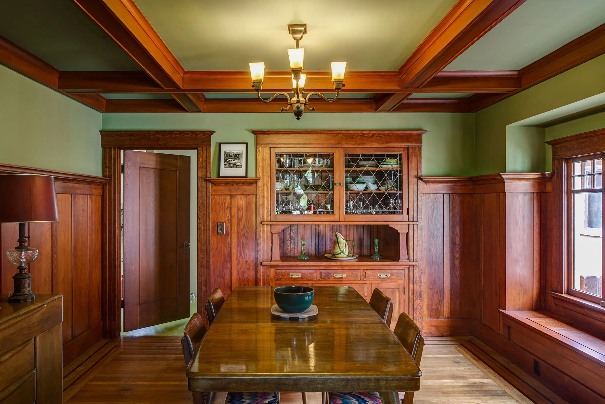 Restored Craftsman dining room with coffered wood ceiling, built-in leaded glass hutch, and a long table, reflecting the home’s original character in the remodel.