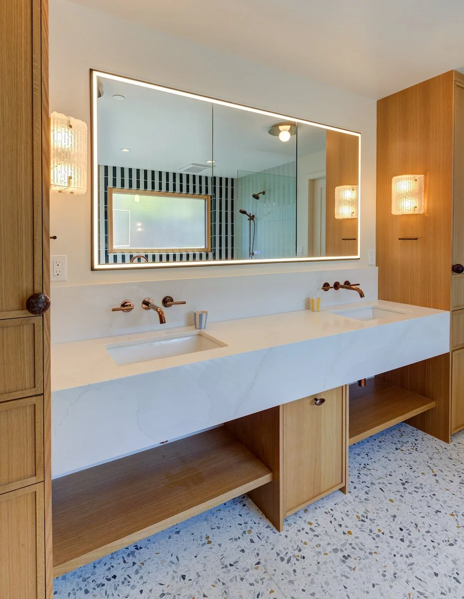 Primary Bath. A white stone countertop is integrated with custom Oak cabinetry and open shelving, creating a warm, streamlined vanity. Backlit mirror and textured sconces add a refined layer of ambient lighting. Terrazzo tiles at floor.
