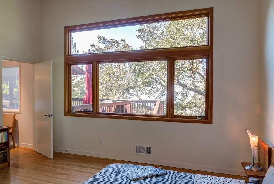 Bedroom created from a former garage, featuring a large wood-framed window with views of trees and nearby deck.