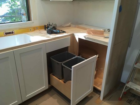 Kitchen base cabinetry under installation featuring integrated pull-out waste and recycling bins beneath the counter near the sink and window.