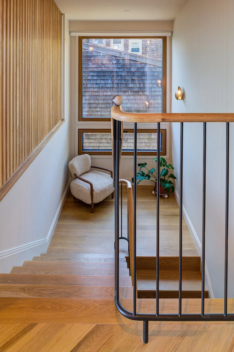 Natural oak treads and a sculptural wood handrail soften the crisp black metal balustrade, creating a refined contrast of warm and industrial materials. The large window at the landing brings natural light into the circulation space.