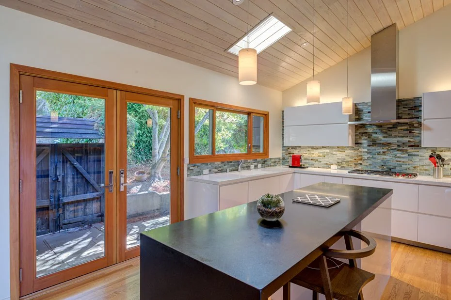 Remodeled kitchen with a central island, white cabinetry, and tiled backsplash, featuring a vaulted wood ceiling with skylight and glass doors opening to a tree-filled outdoor area.