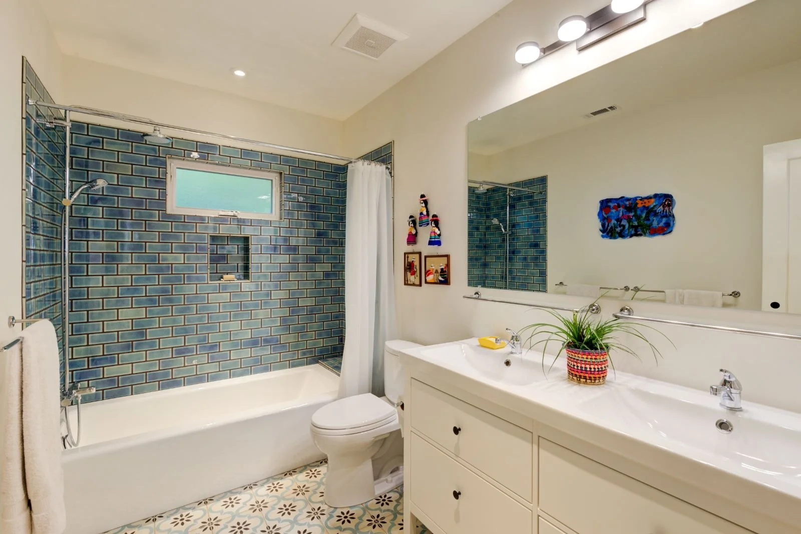 New Bathroom with dark blue and teal tiles at the tub/shower surround contrast with the white walls, vanity, bathtub, and patterned floor tiles. Double sinks with a long mirror. 