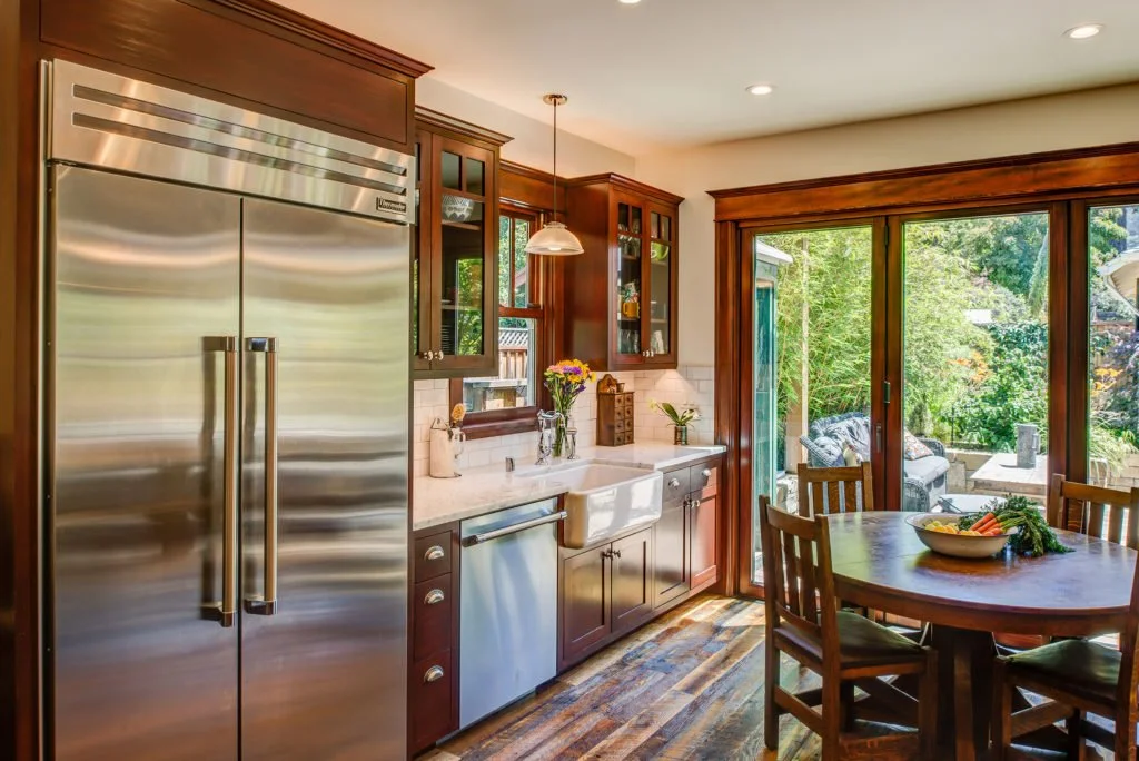 View past the refrigerator and tall pantry cabinets toward the kitchen and exterior folding doors, showing the open plan connection from kitchen to the deck.