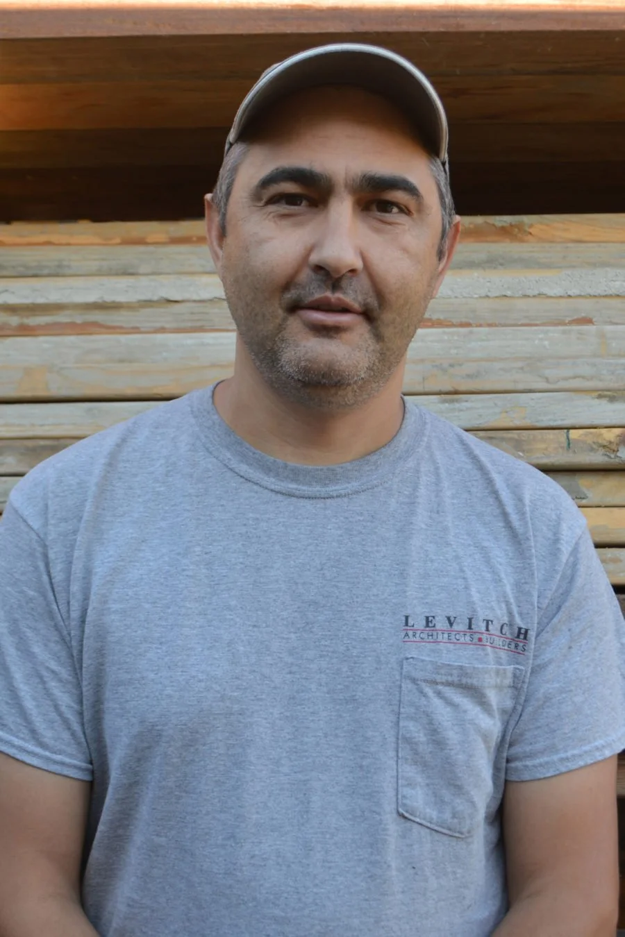 A man wearing a gray Levitich Architects t-shirt and a baseball cap, standing outdoors in front of a wooden fence.