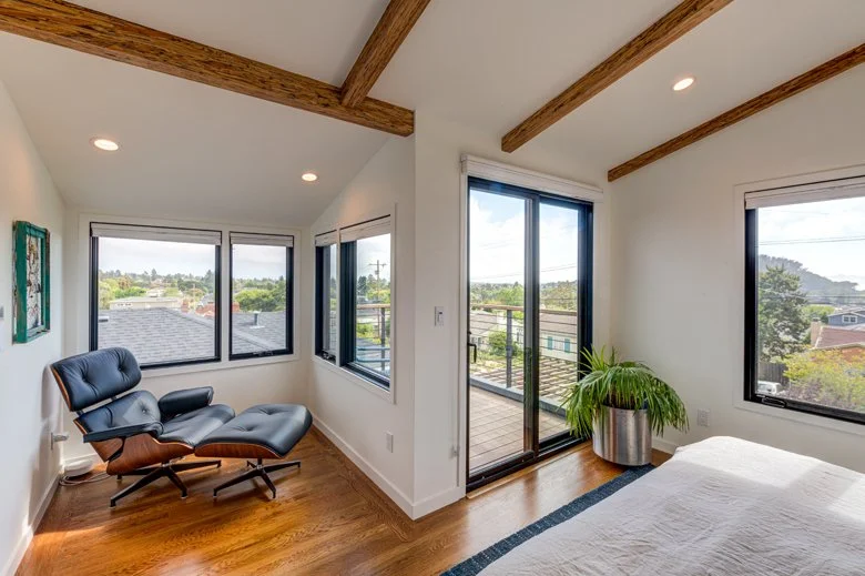 Primary bedroom with exposed wood ceiling beams, wide windows, and sliding glass doors opening to a private balcony, creating a light-filled retreat with views over the surrounding neighborhood.