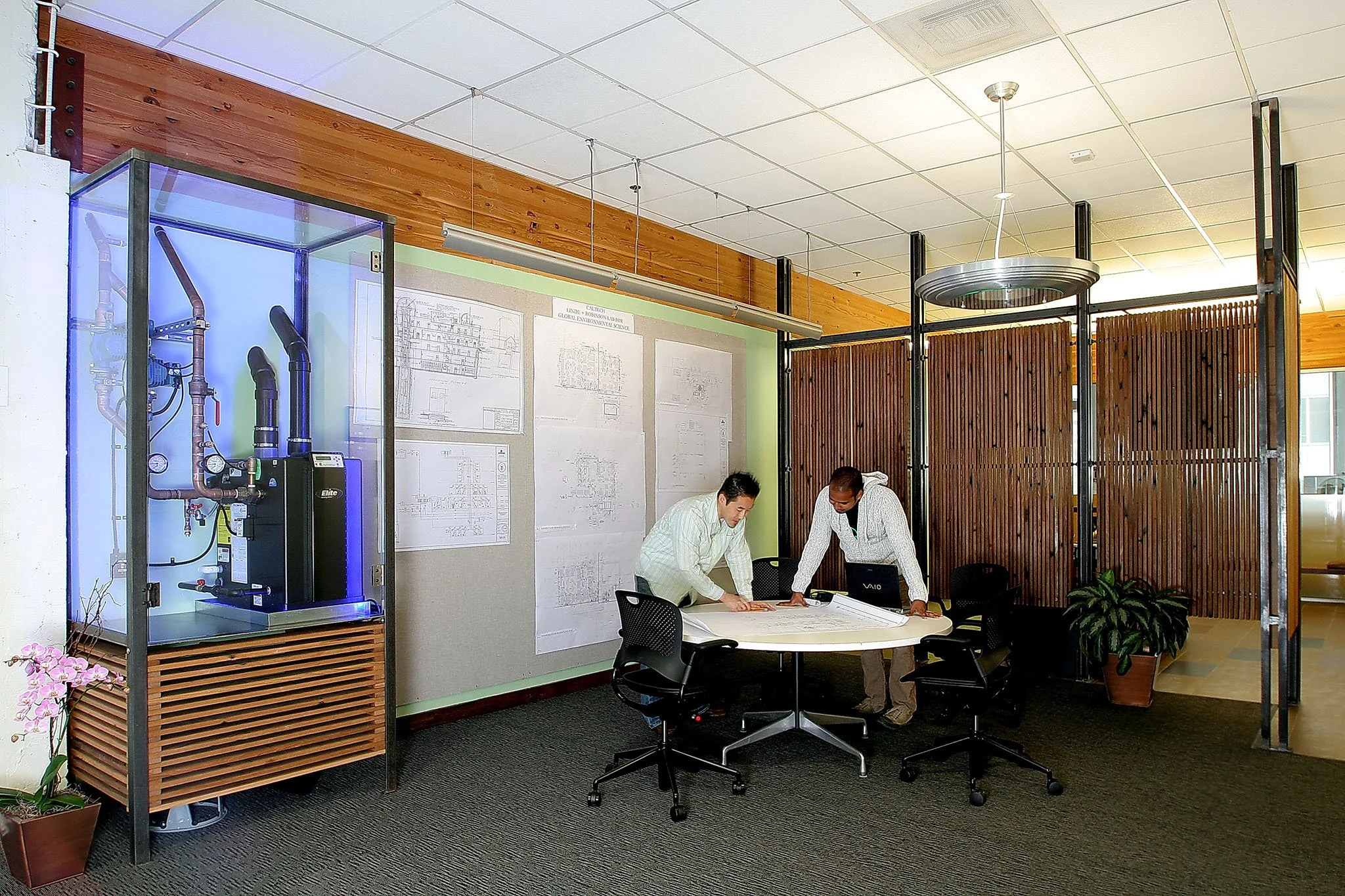 Collaborative meeting area with daylighting, recycled wood screens, and a display case showing the hydronic heating boiler and piping while staff review plans at a round table.