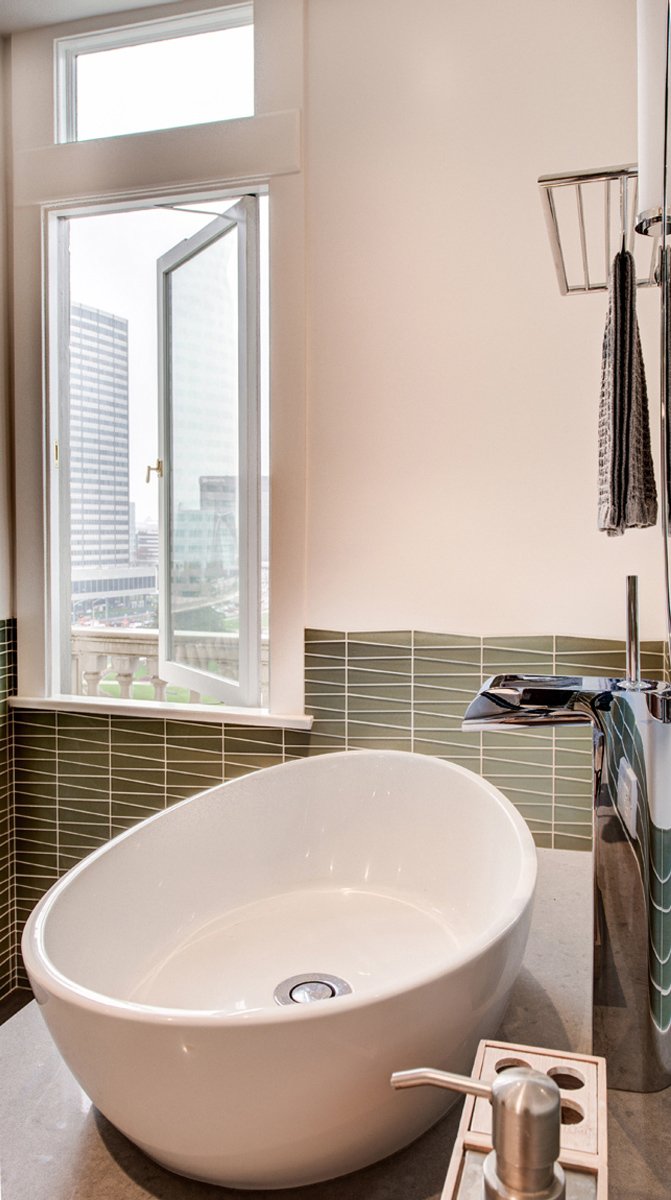 Freestanding oval soaking sink beneath a window with city views, framed by green glass tile wainscoting in the expanded primary bathroom.