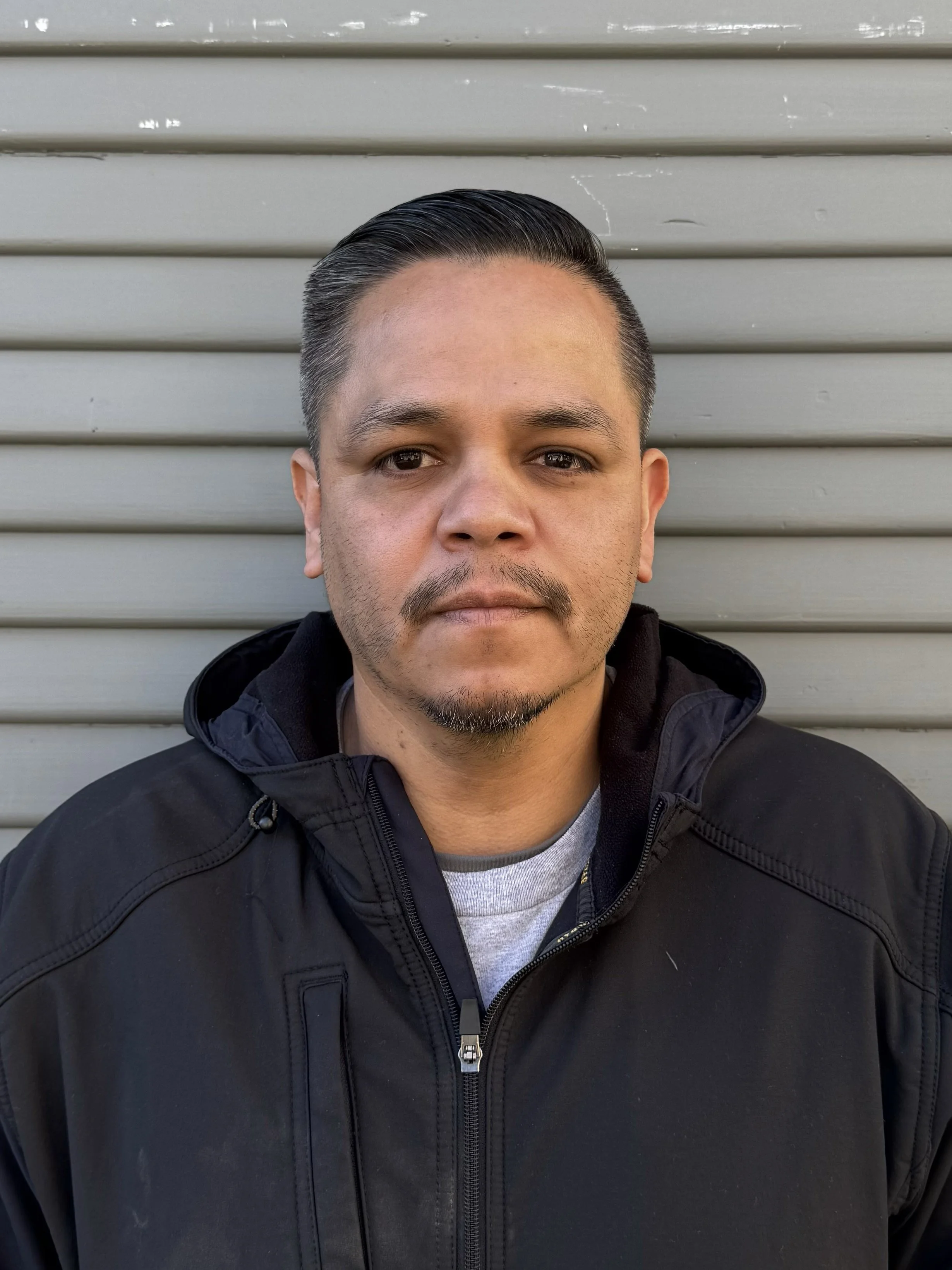 A man with short, dark hair and a goatee, wearing a black jacket, standing in front of a gray, horizontal metal siding background.